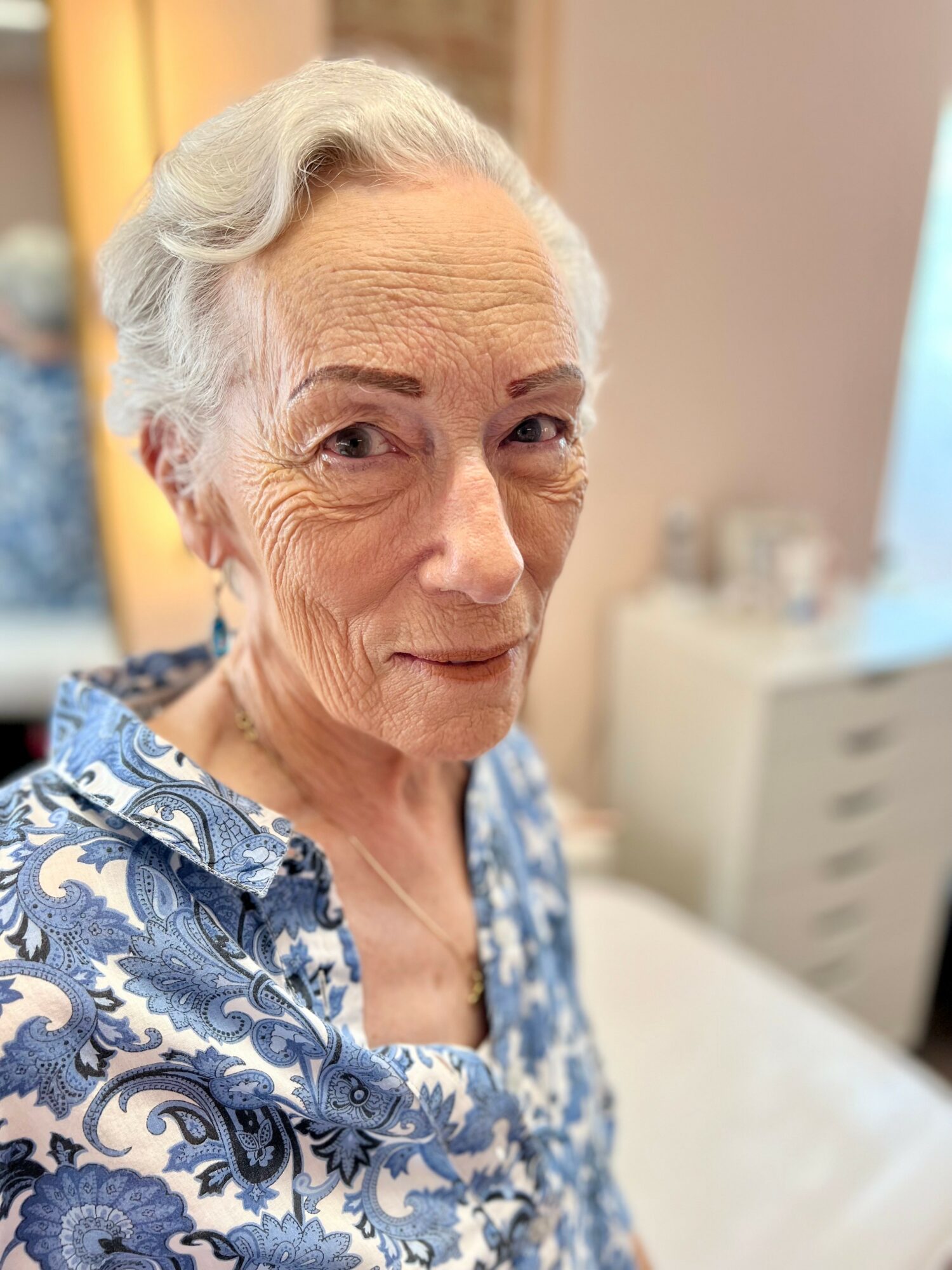 Elderly woman with gray hair and a patterned blue and white blouse, smiling softly indoors.