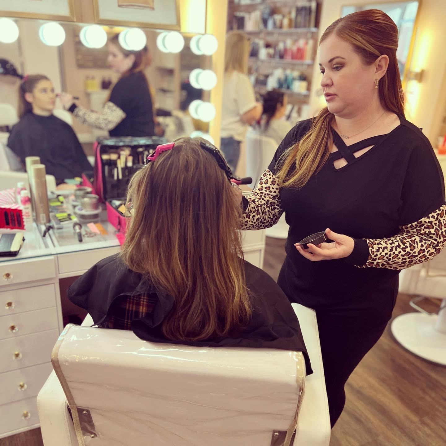 Makeup artist applying makeup to a woman seated in a salon chair, with mirrors and beauty products in the background.