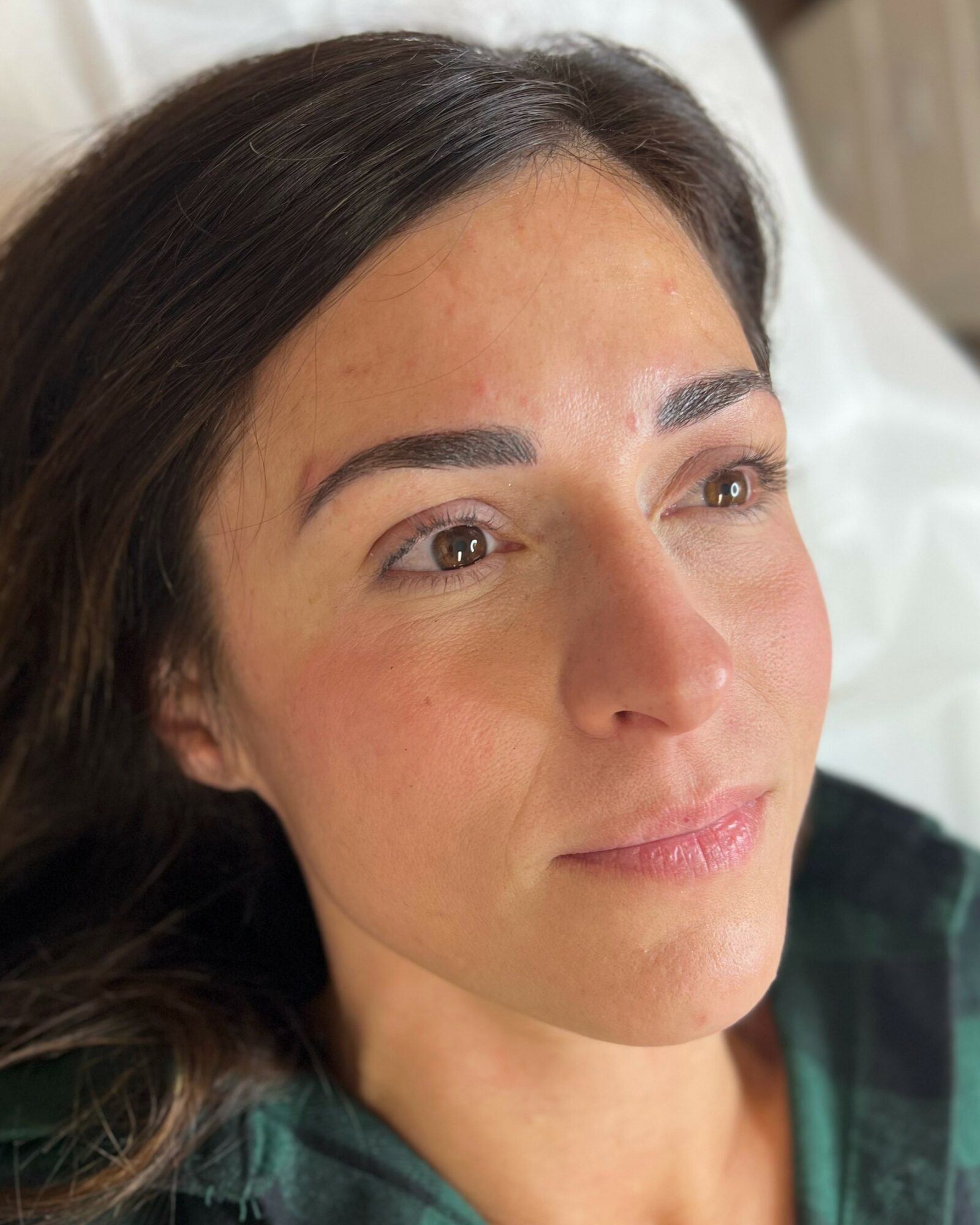 Close-up of a woman with dark hair lying on a bed, looking to the side, with natural light on her face.