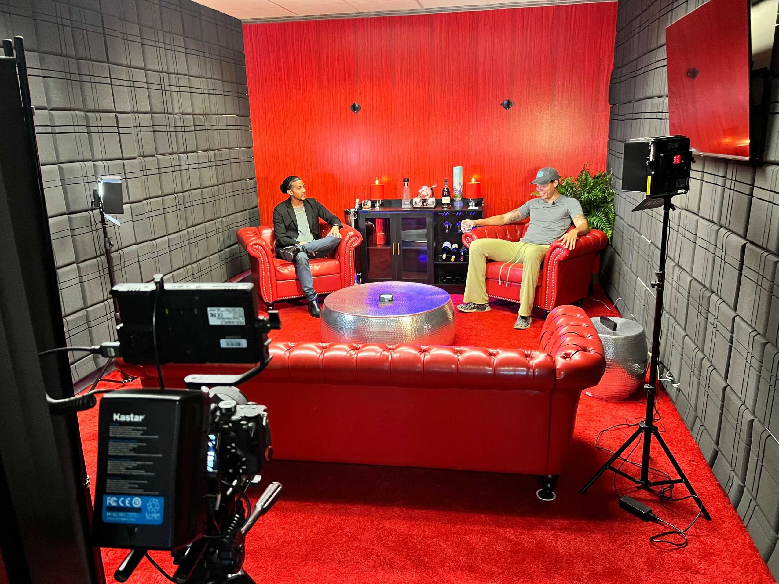 Two people sit on red leather chairs in a recording studio with black soundproof walls and a red background, with equipment around.