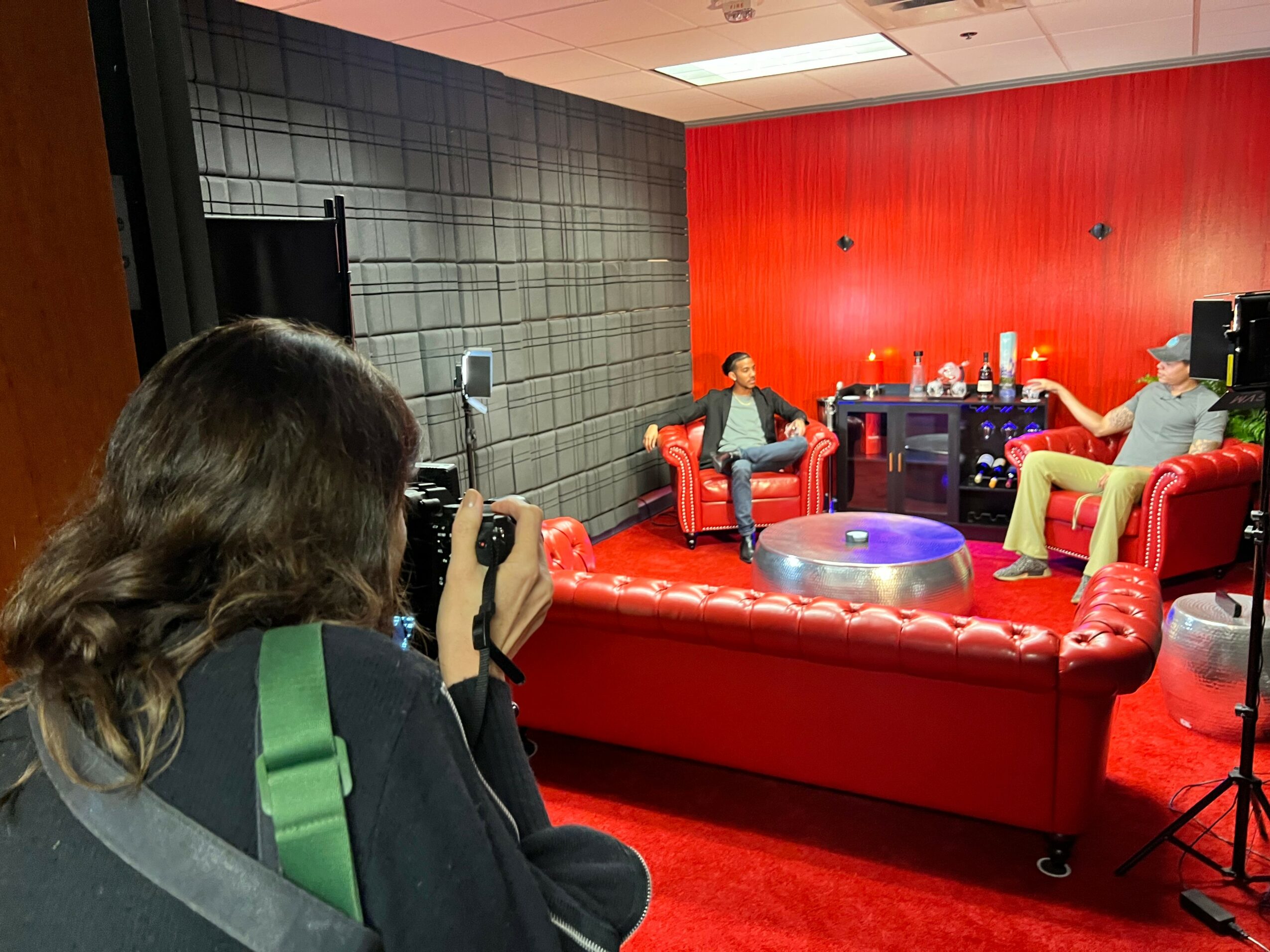 Person with long hair taking a photo of two people sitting on red couches in a room with red and black walls.
