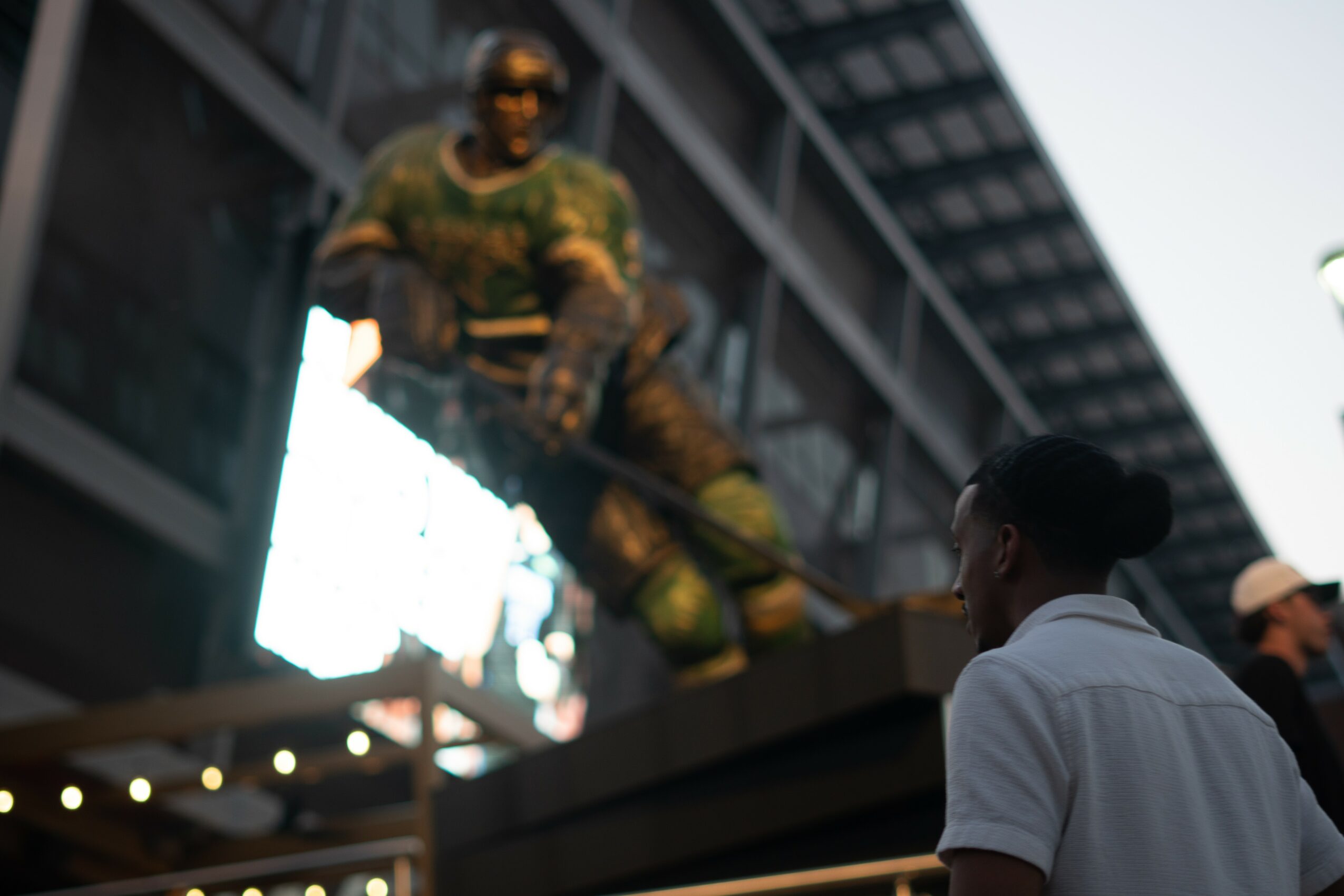 Person with dark hair in a white shirt looking at a large statue of a soldier holding a rifle, outside a building.