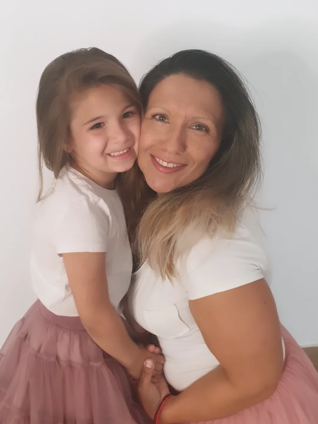A woman and a young girl smiling, standing close together against a plain white background.