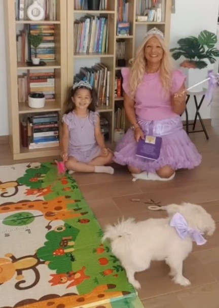A woman and a girl sit on the floor in a room with bookshelves, a colorful rug, and a white dog with a bow.