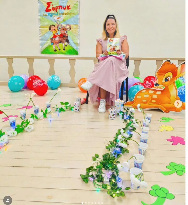 Woman sitting on a chair holding a book, decorated with balloons, flowers, and a large cartoon animal figure, in a festive setting.