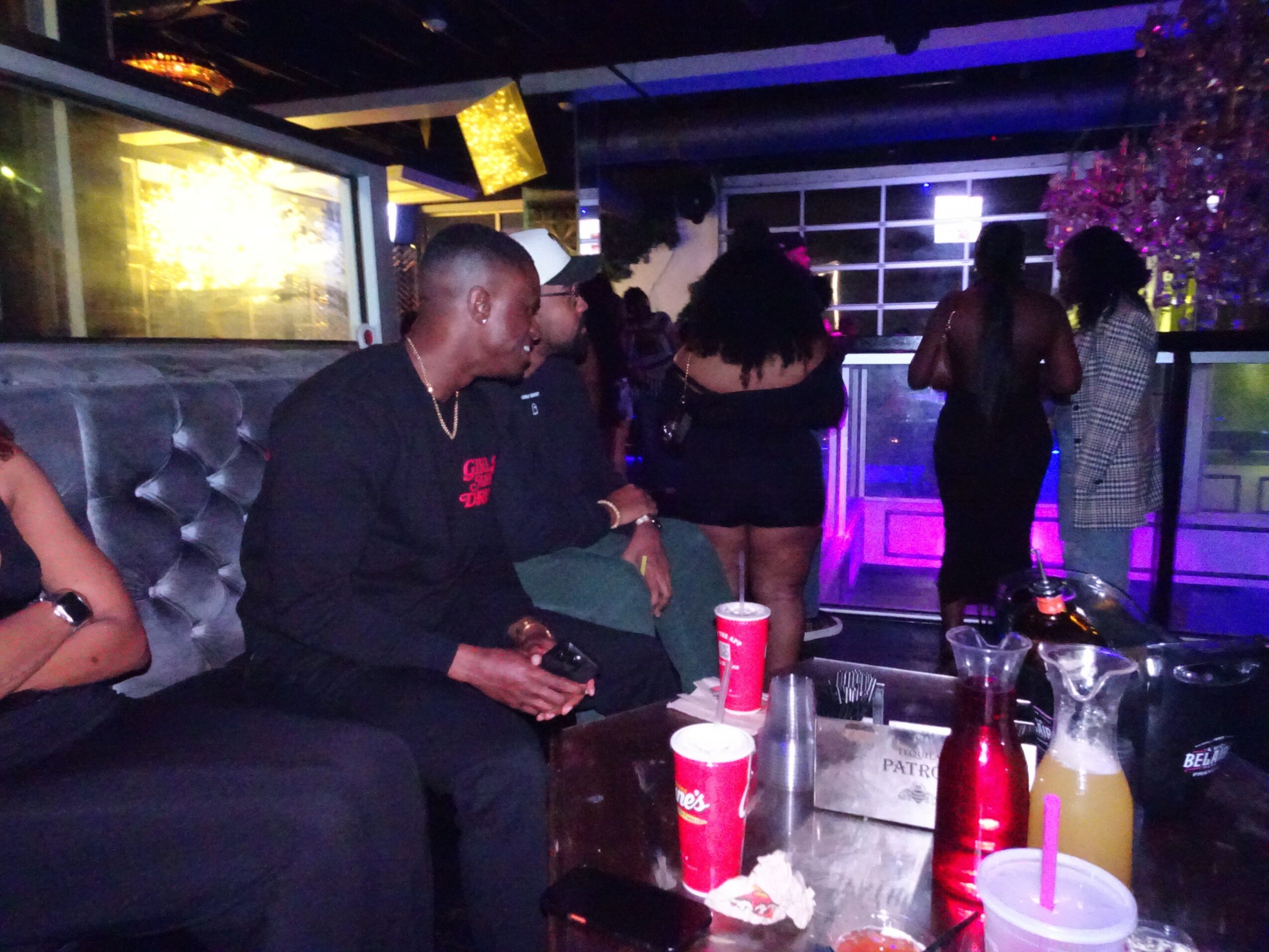 People socializing in a bar with drinks and colorful lighting, some standing and some sitting, in a lively setting.