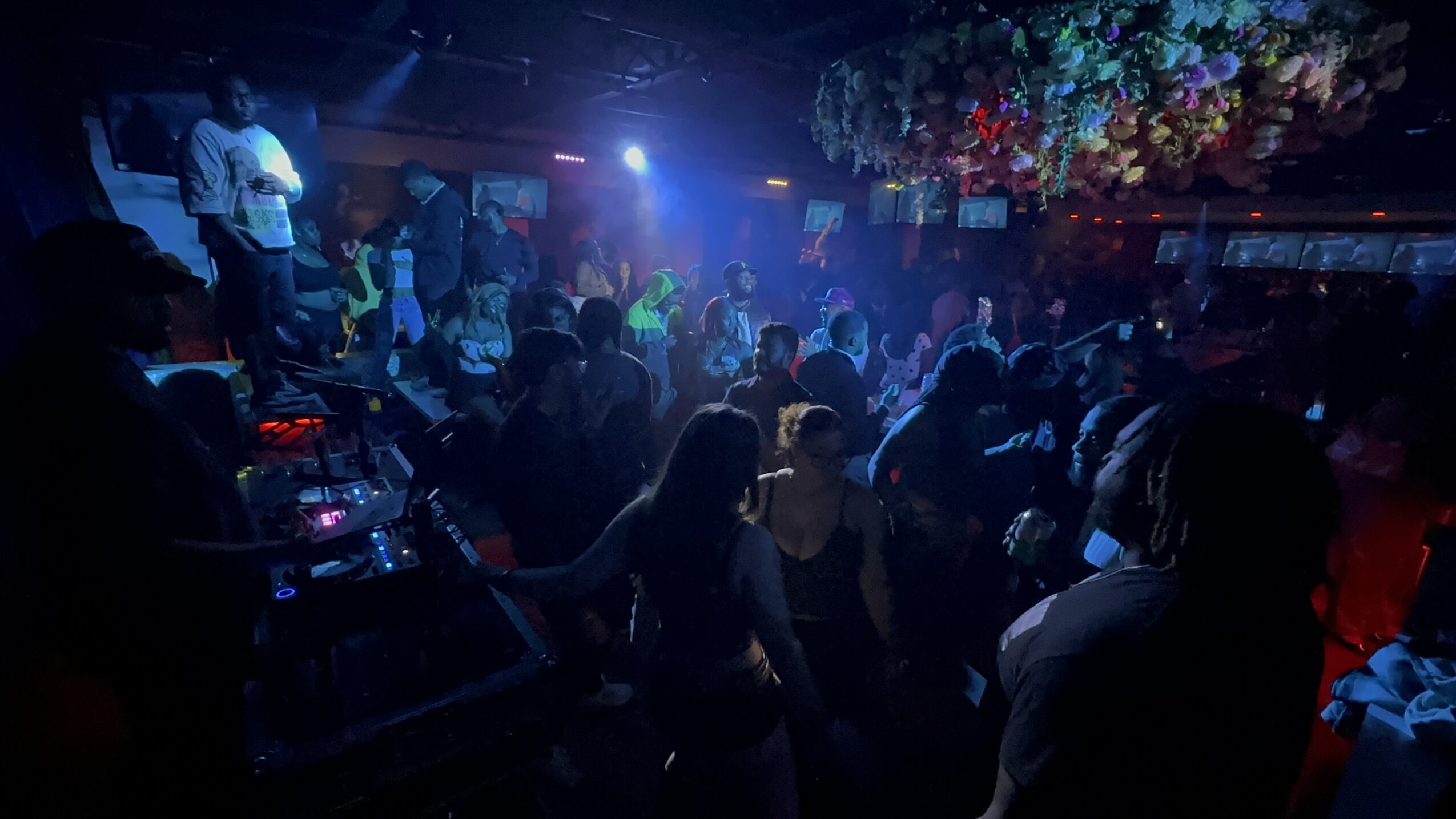 Crowded nightclub with people dancing under colorful lights and hanging decorations, dark atmosphere, lively scene.