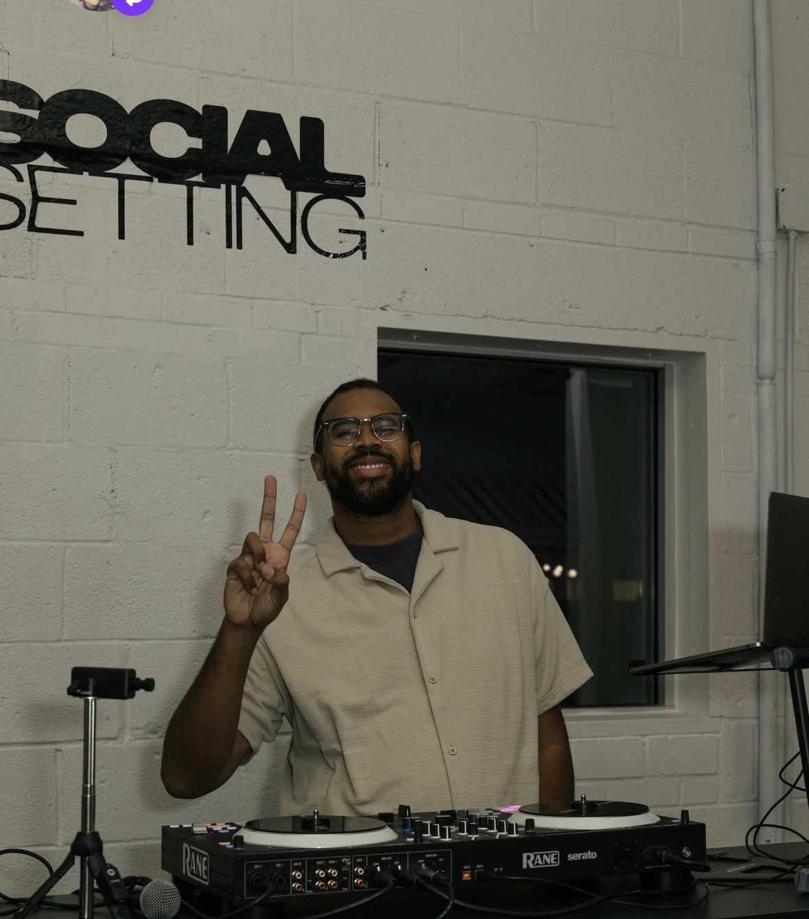 Man with glasses and beard smiling and making a peace sign, standing in front of a white wall with a window and equipment.