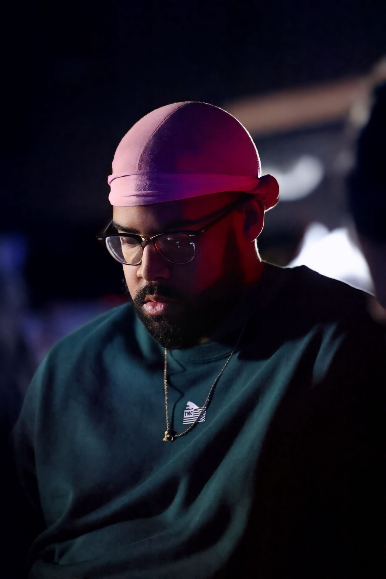 Person wearing glasses and a pink headscarf, looking down in a dark setting.