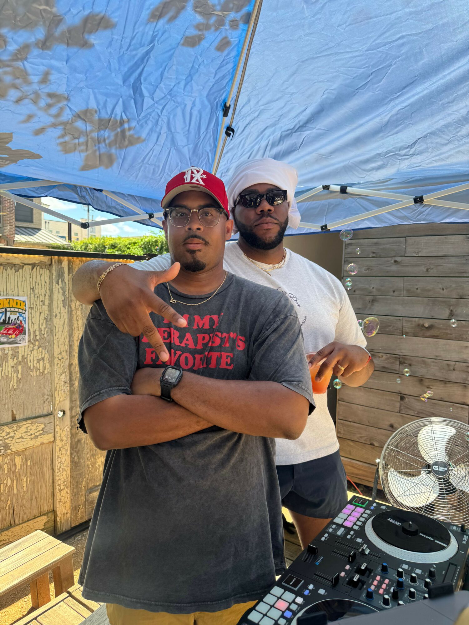 Two men standing outdoors near a wooden fence, one wearing sunglasses and a white cap, the other in a red cap, both making hand gestures.