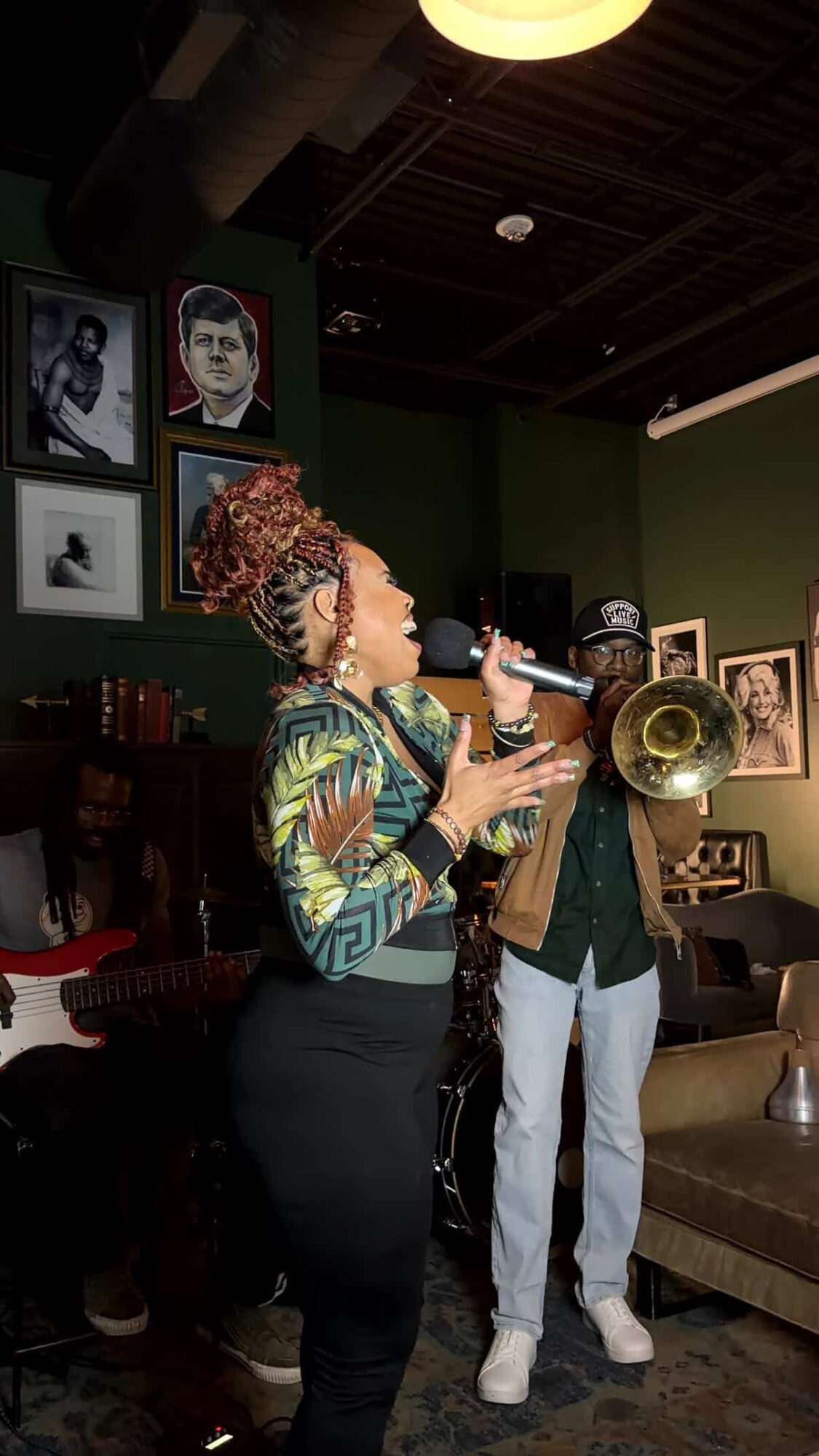 Woman singing into a microphone and playing a trumpet in a room with framed photos on the wall.