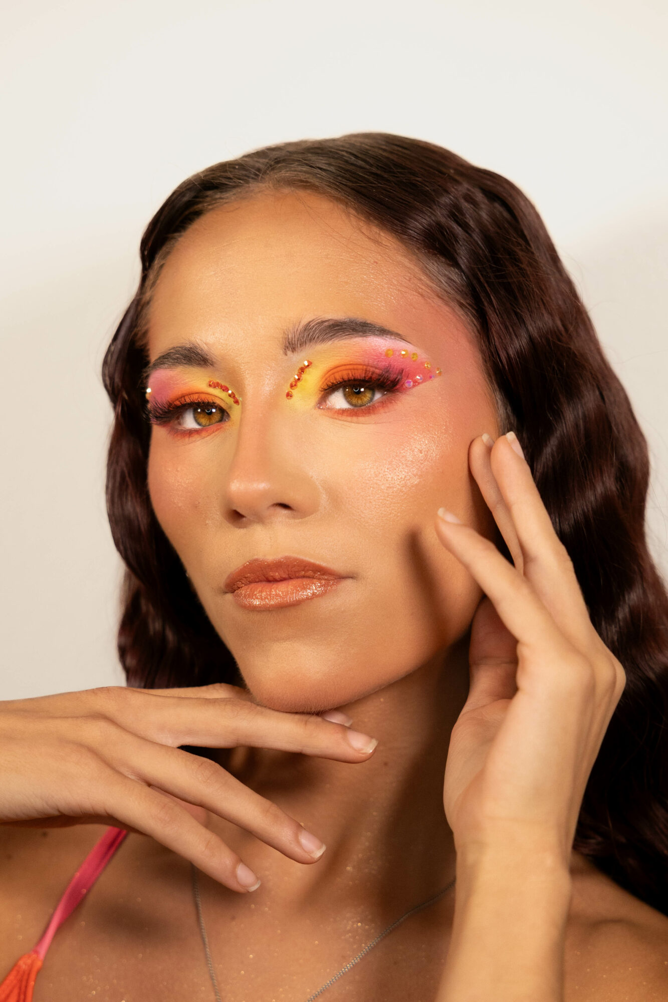 Woman with colorful eye makeup and wavy hair, touching her face gently, against a plain background.