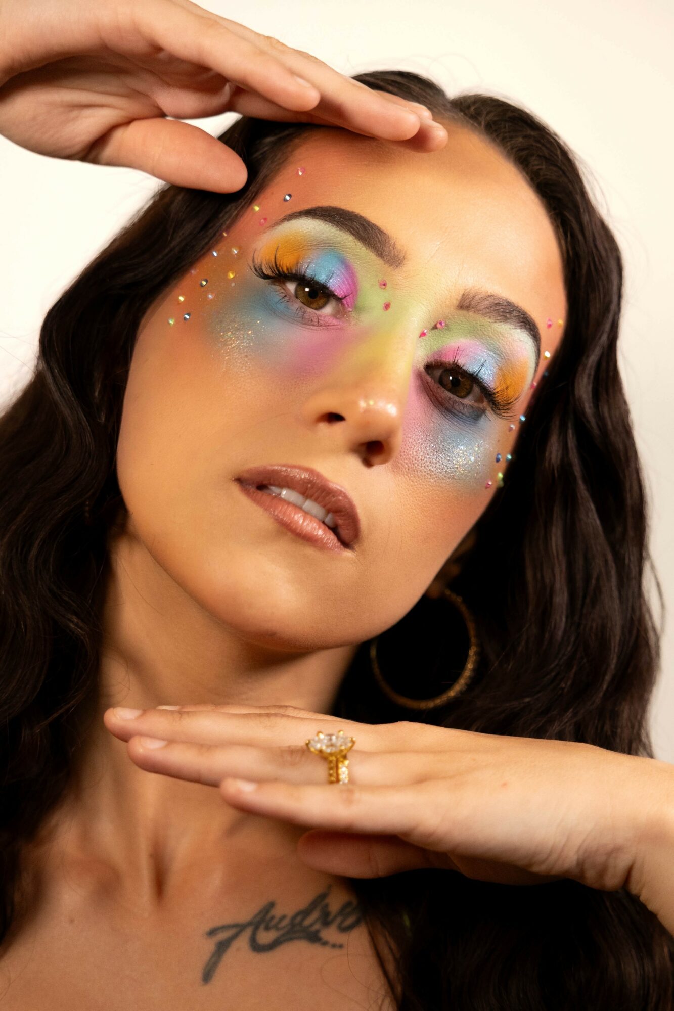 Woman with colorful rainbow makeup and star-shaped glitter, posing with hands near face, dark hair, hoop earrings, and a tattoo.