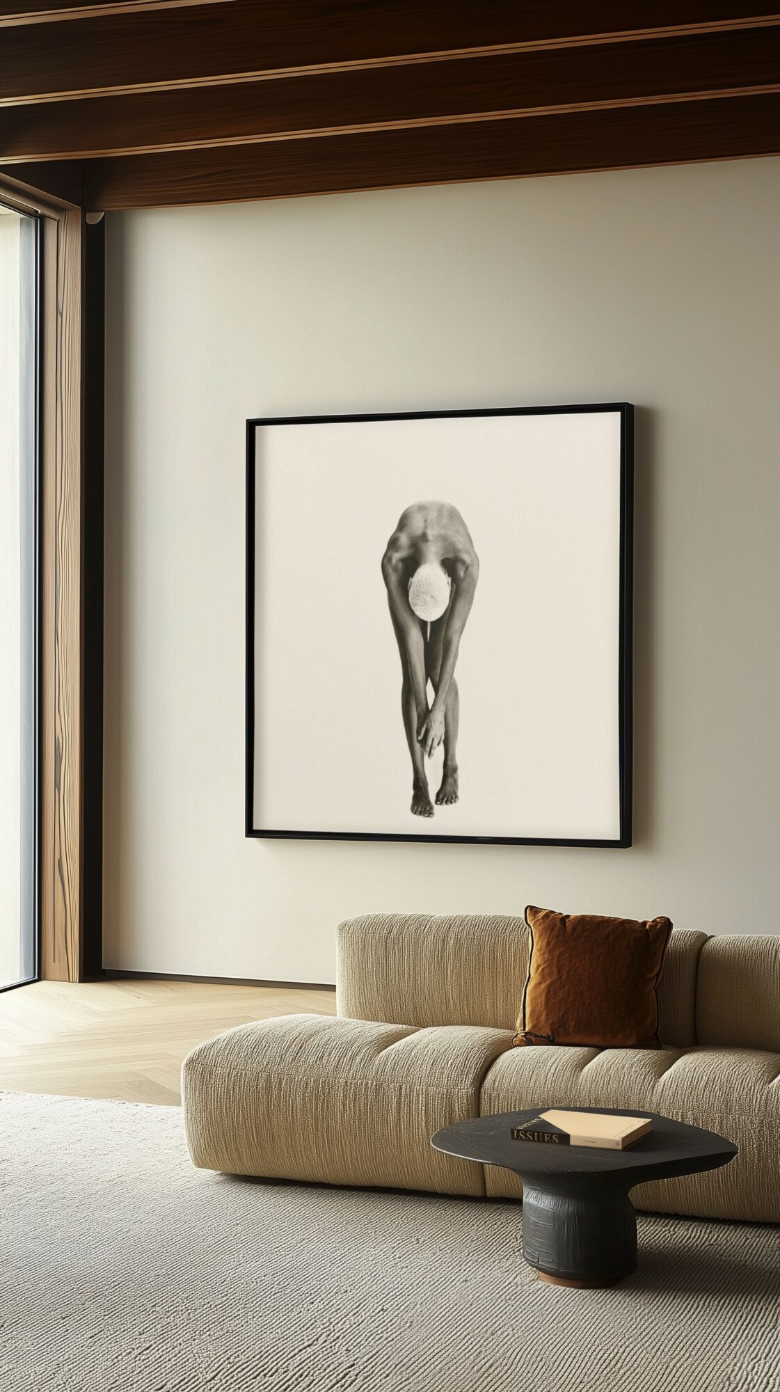 Living room with a framed black-and-white artwork of a person bending over, beige sofa with a brown pillow, and a small black coffee table.