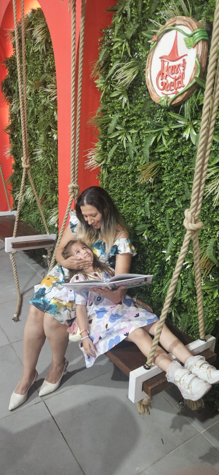 Two children sit on a swing with a woman, surrounded by green plants and holiday decorations, including a sign with a star.