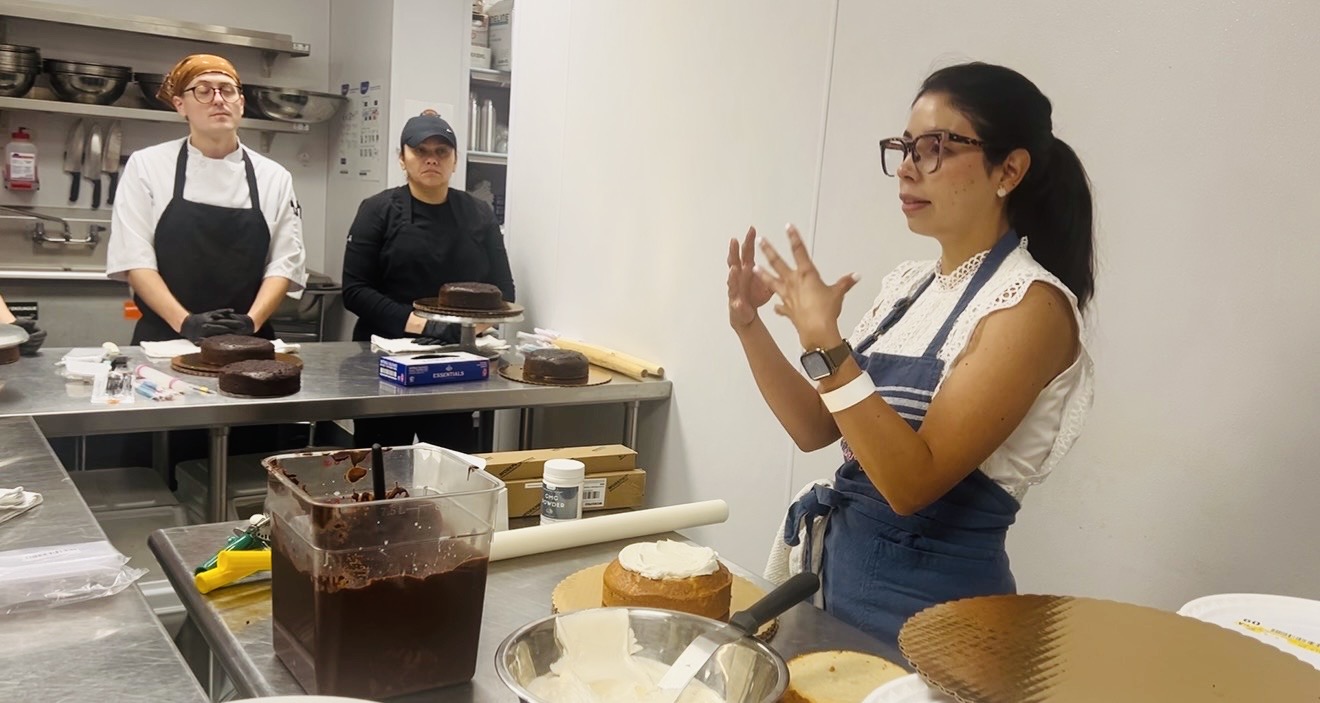 Woman in apron giving a cooking class, three people observing in a kitchen, various cooking tools and ingredients visible.