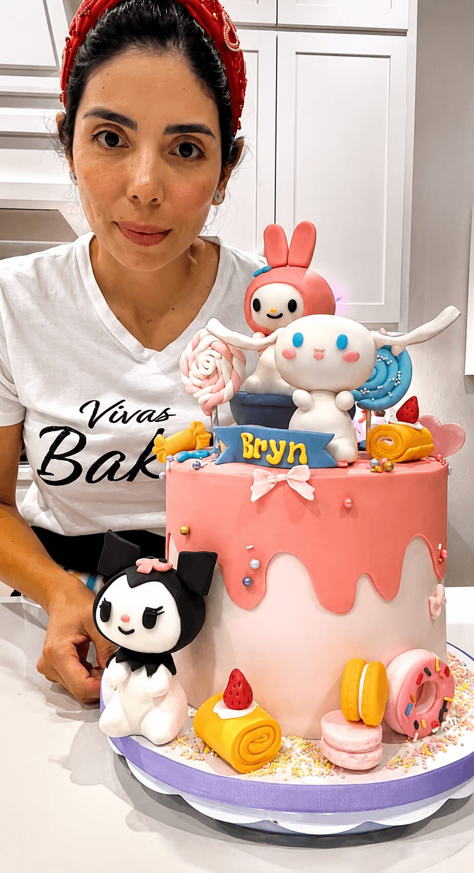 Woman with dark hair and red headband behind a pink cake decorated with cartoon characters and sweets, in a kitchen.
