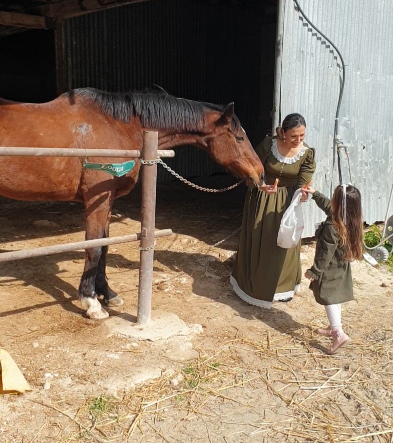 Woman and girl interact with a brown horse in a farm setting, woman holding the horse's bridle, girl reaching out.