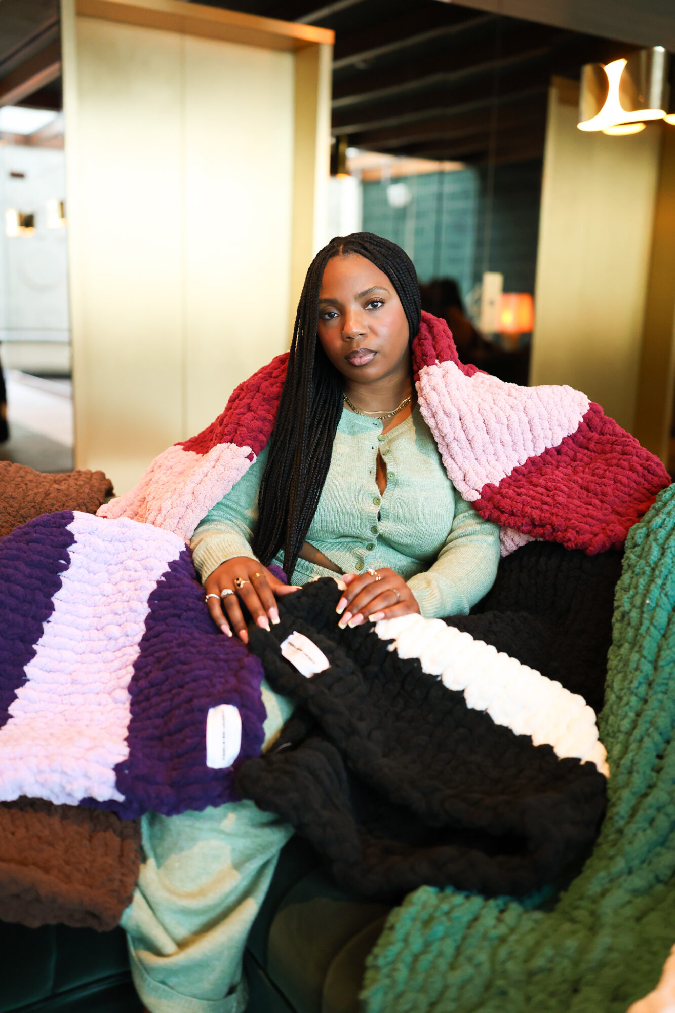 Woman with long black hair sitting on a couch, wearing a light green outfit, surrounded by colorful knitted blankets.