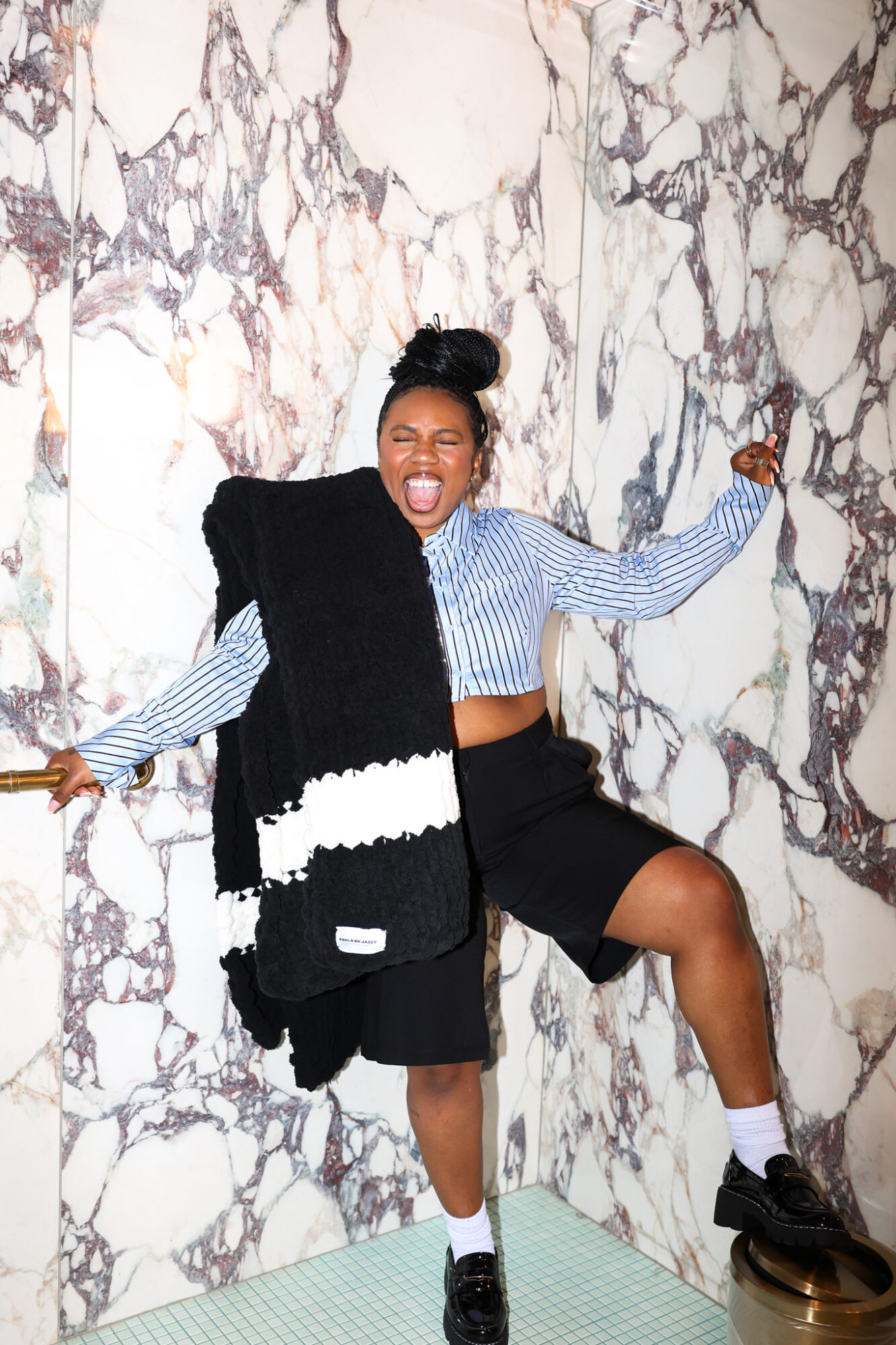 Young woman laughing with arms outstretched, standing against a marble wall, wearing a striped shirt, black shorts, and sneakers.