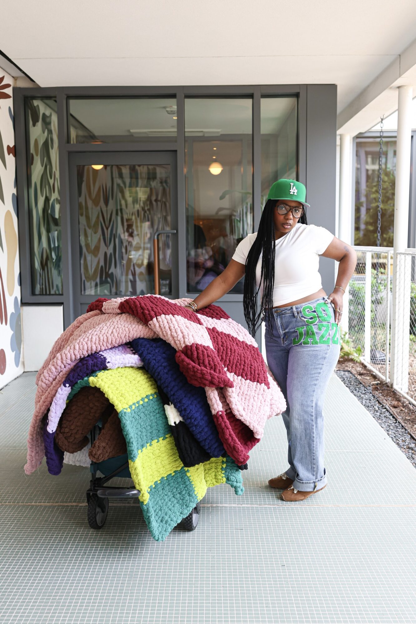 Woman with long hair, wearing a green cap, white shirt, and jeans, stands next to a cart of colorful knitted blankets outside a building.