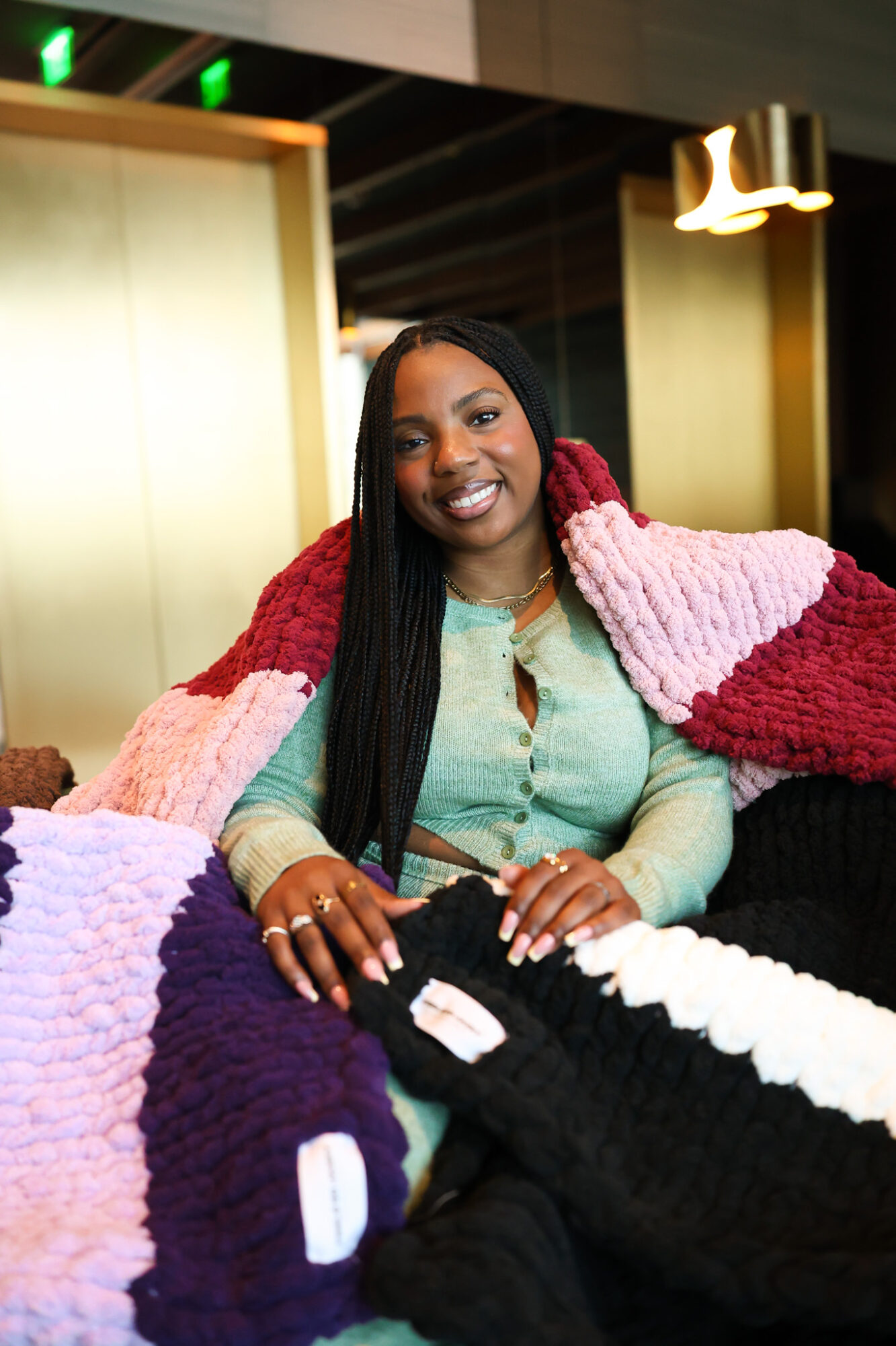 Smiling woman with long black hair wearing a pastel green top and a multicolored knitted blanket, sitting indoors.