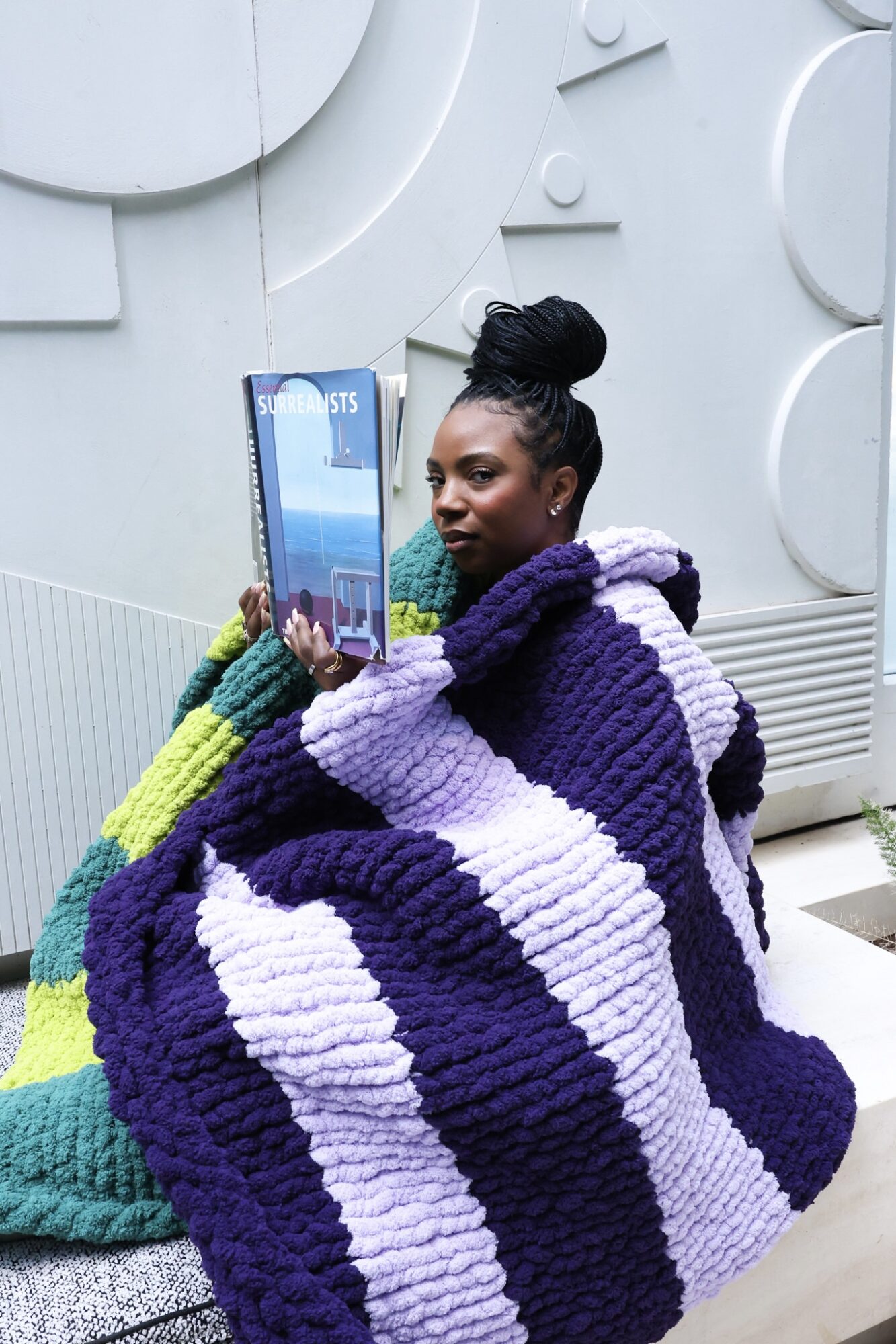 Woman wrapped in a large purple and white striped blanket, holding a tablet, sitting against a white wall.