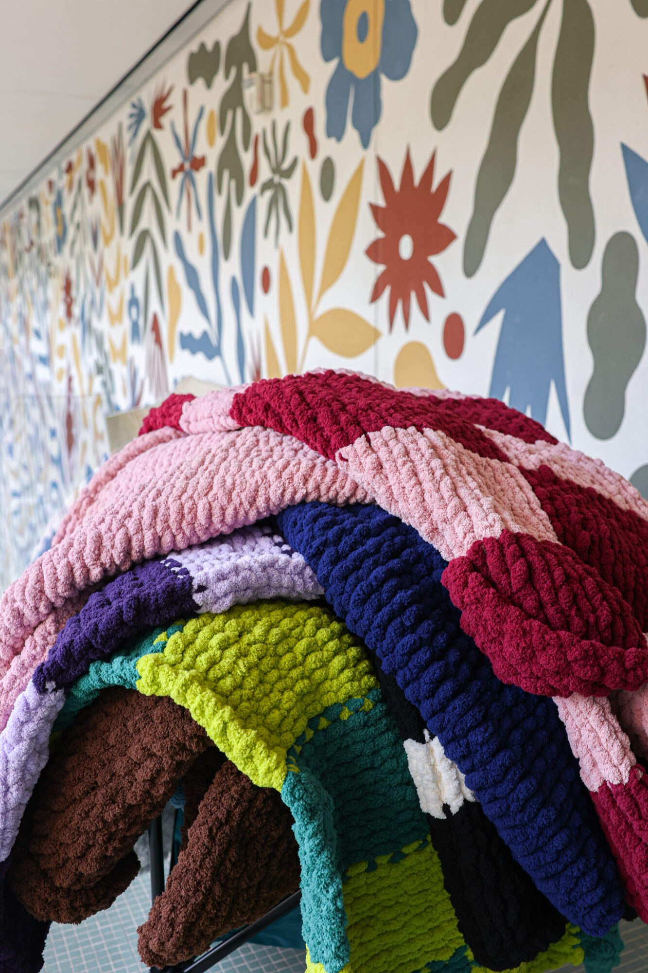 Colorful towels stacked in front of a floral patterned wall, with various colors and textures visible.