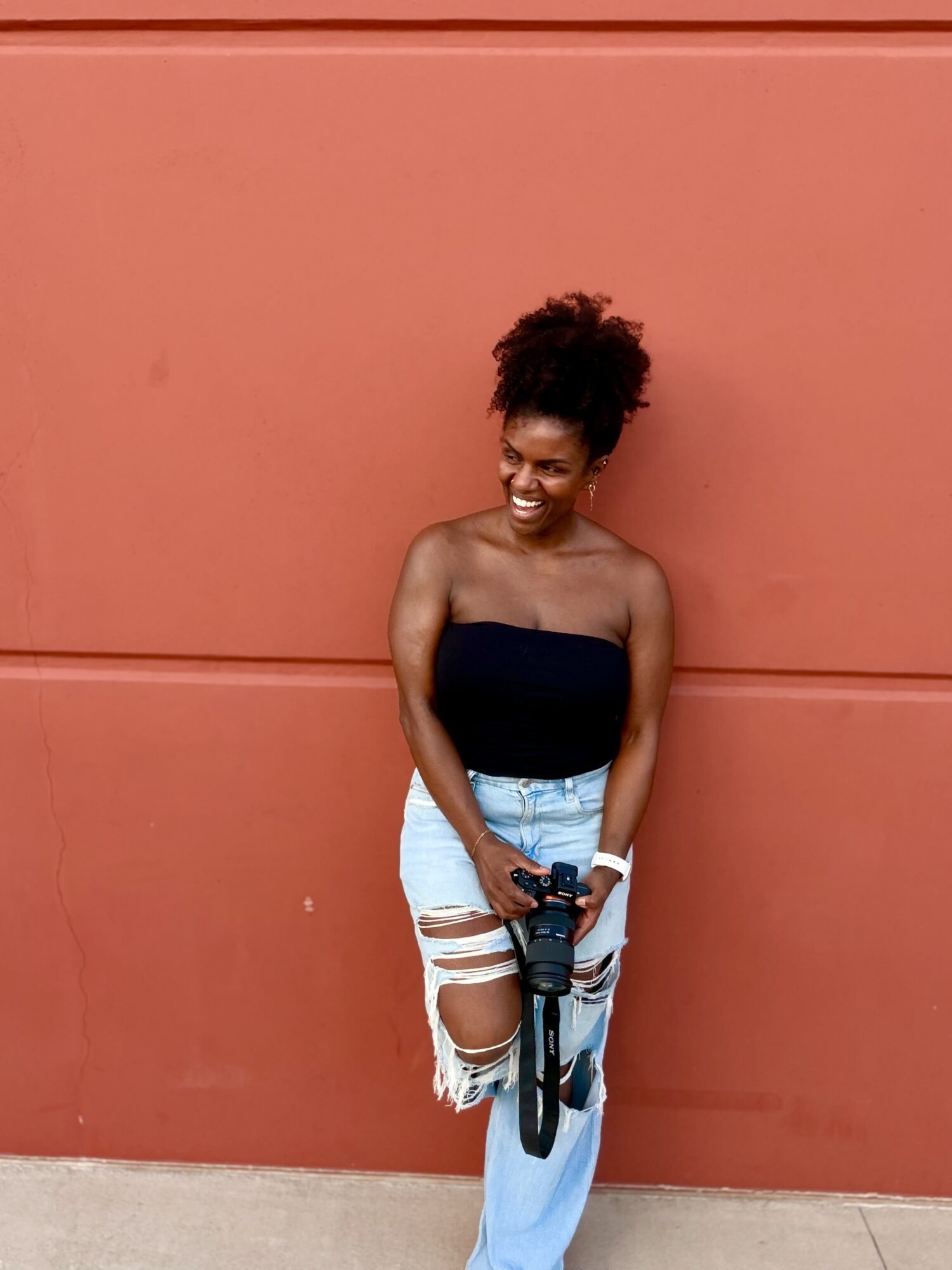 Smiling woman with natural hair, wearing a strapless top and ripped jeans, holding a camera, standing against a red wall.