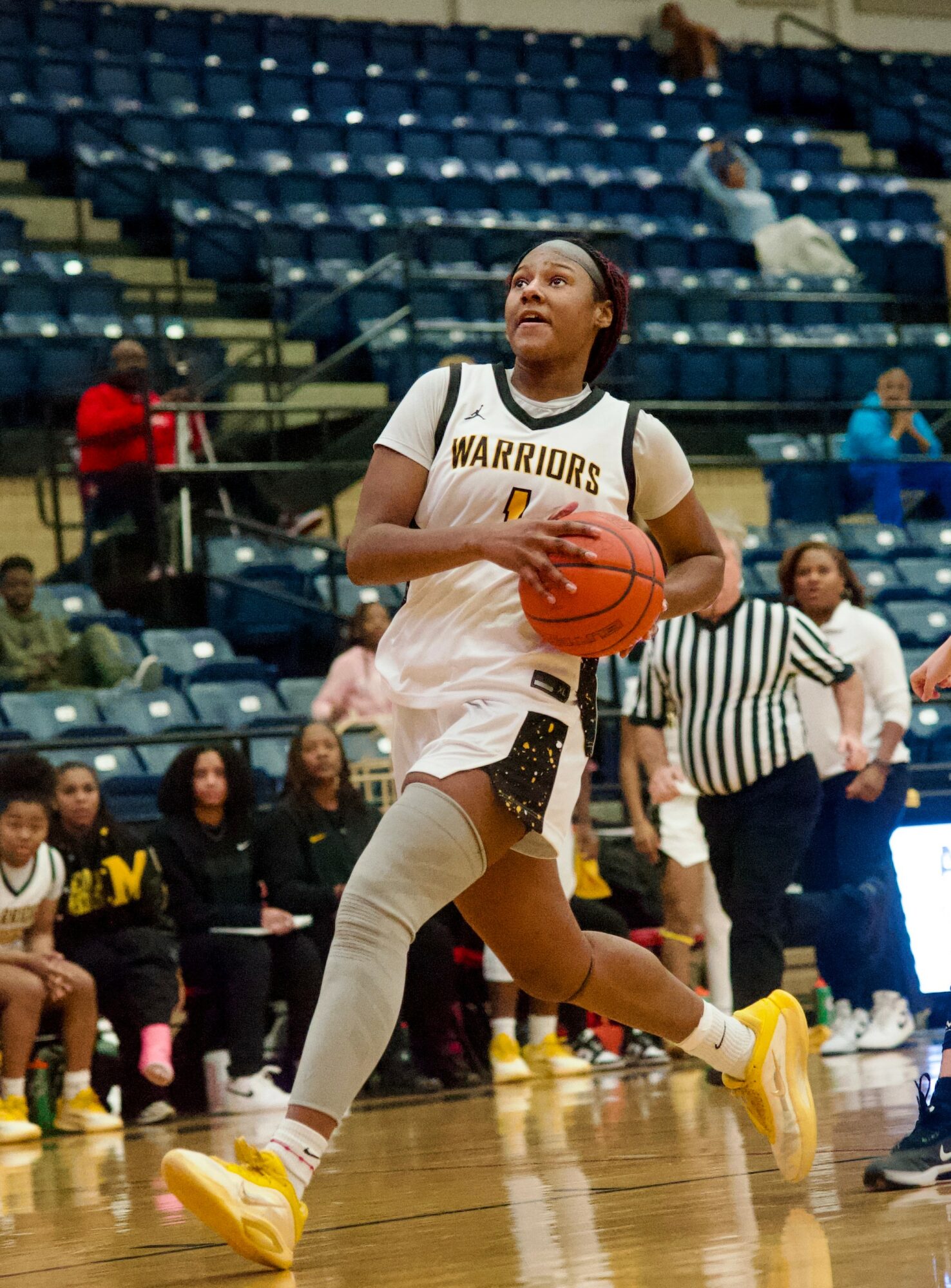 Female basketball player in a white uniform dribbling on the court with spectators and a referee in the background.