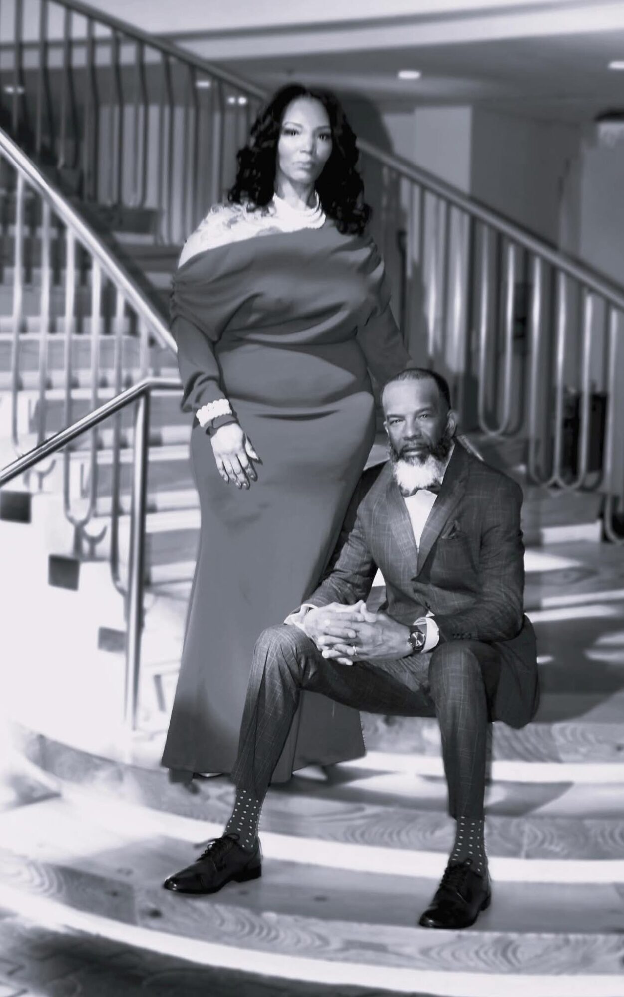 A woman in an elegant dress stands beside a seated man in a suit on a staircase.
