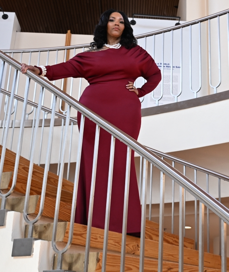 Woman in a long red dress standing on a staircase railing, hand on hip, looking to the side.
