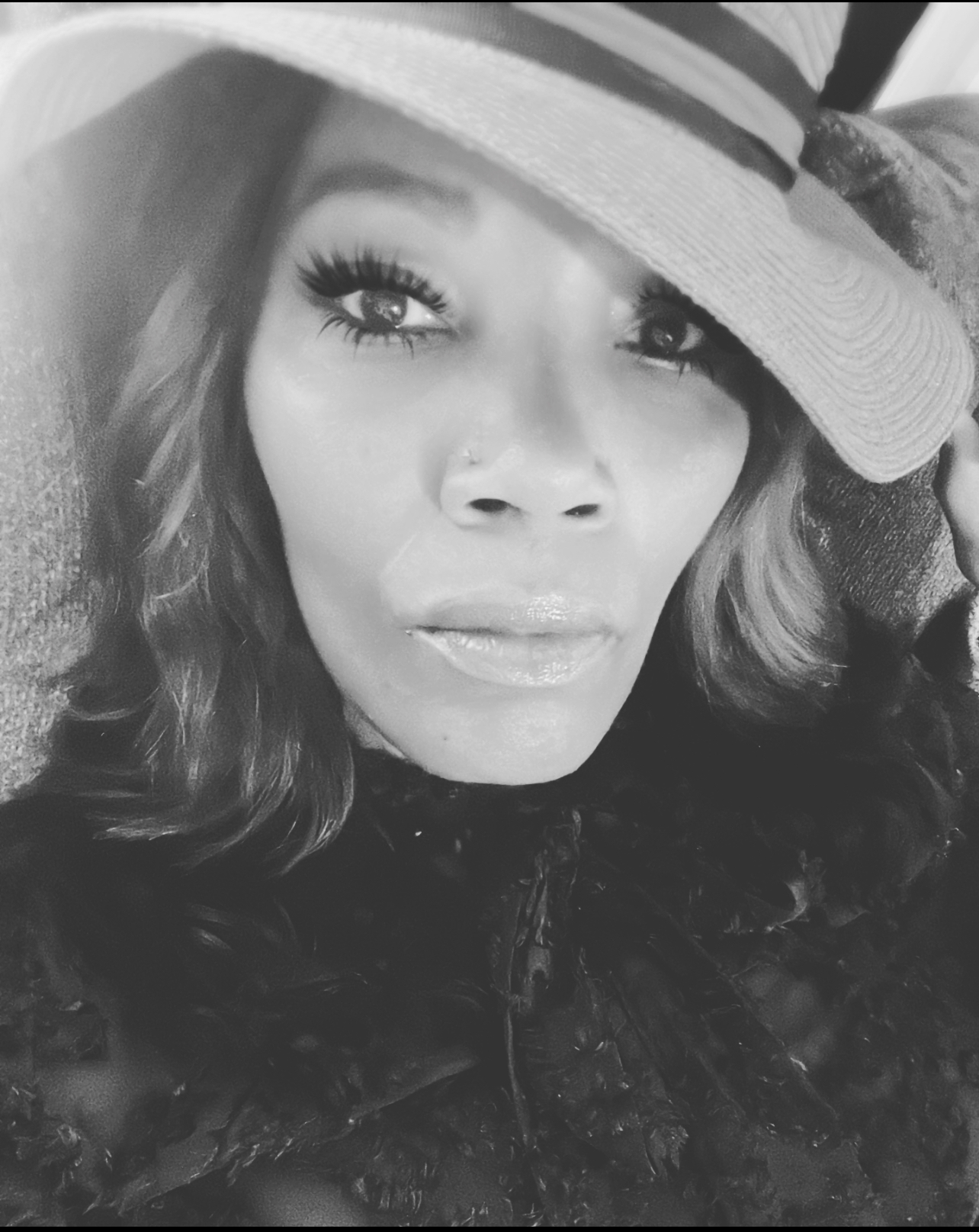 Woman wearing a hat with a serious expression, long wavy hair, and makeup, in black and white.