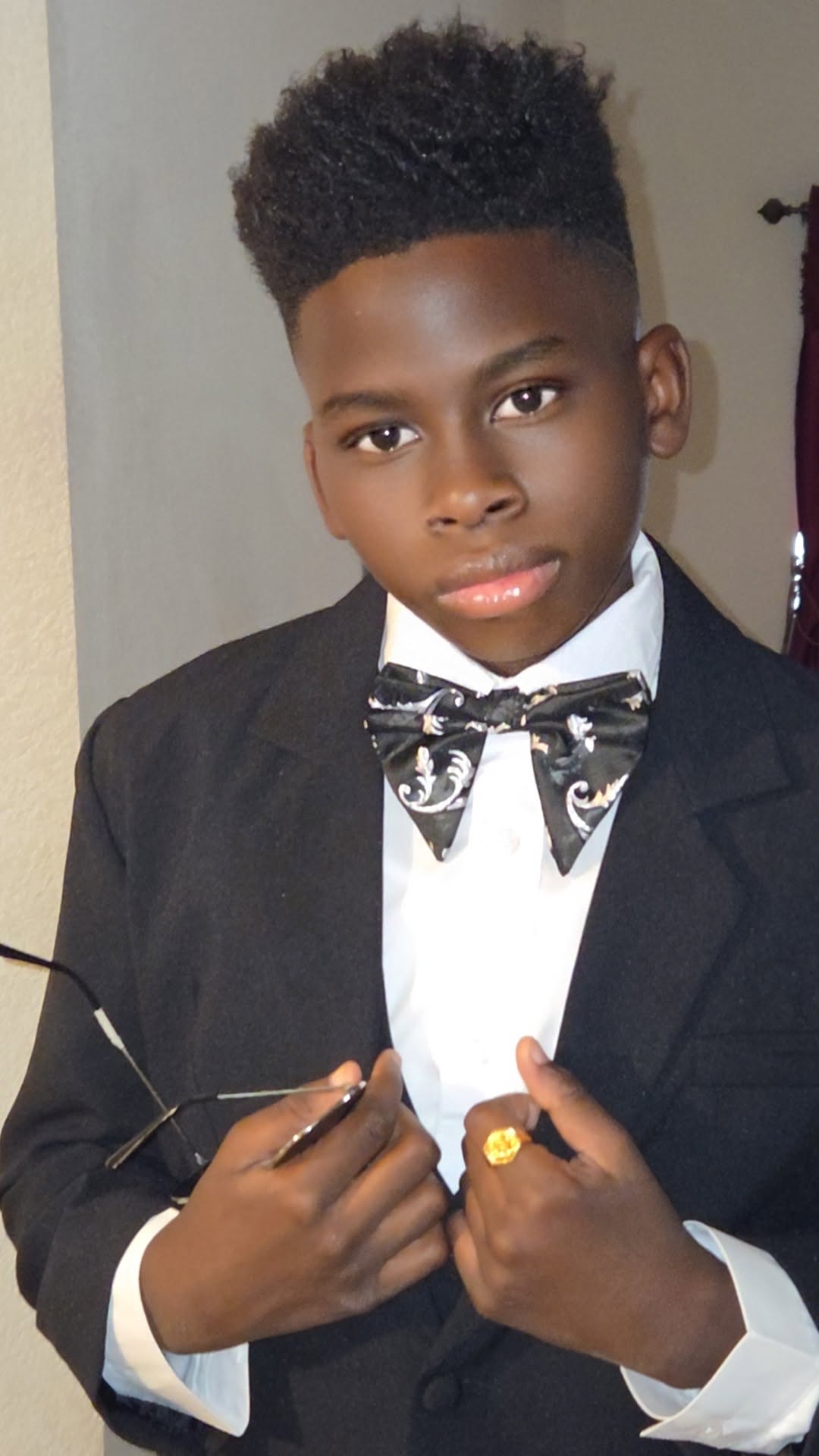 Young boy in formal suit with bow tie, holding glasses and wearing a ring, looking at the camera.