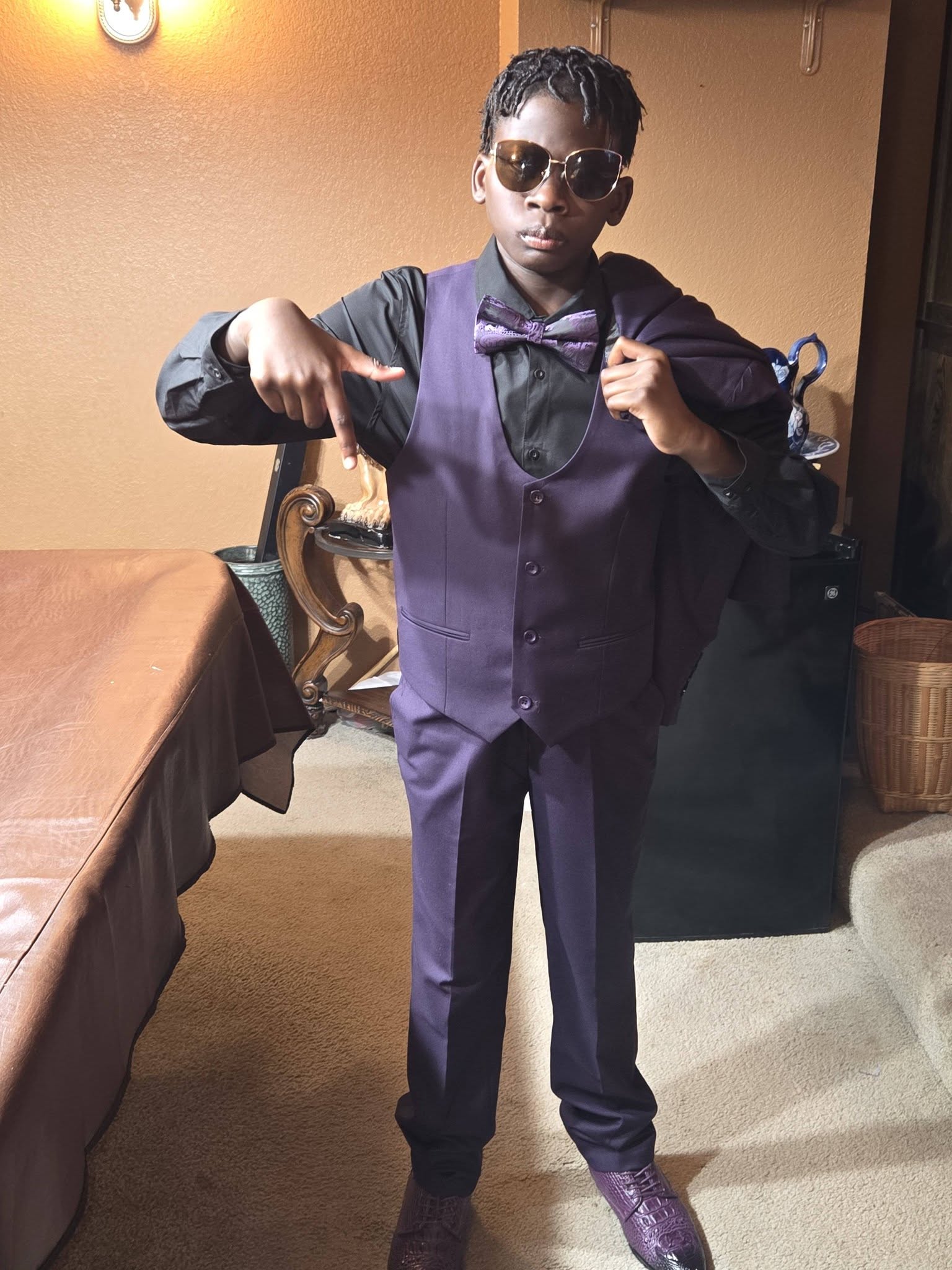 Child in a purple suit, sunglasses, pointing at themselves, standing indoors near a table and a basket.