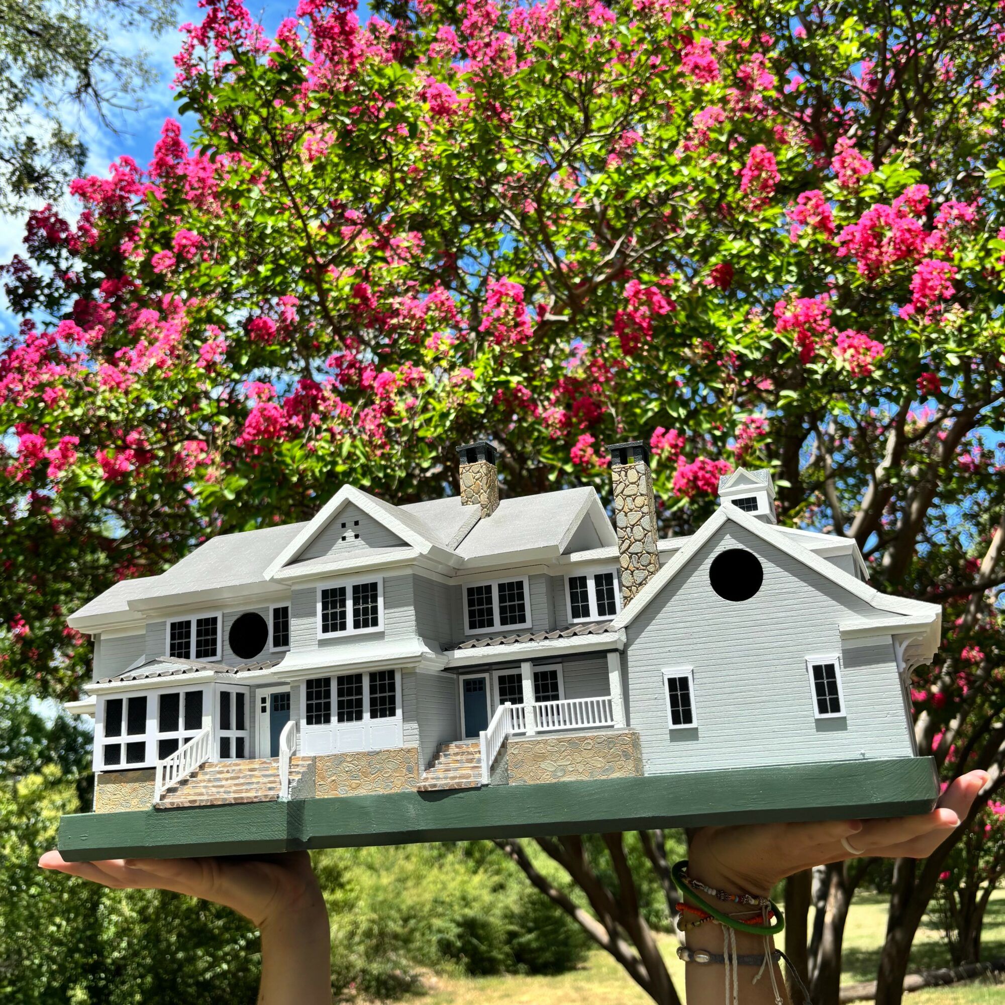 A detailed model of a large white house with multiple levels and porches, held above green grass with a flowering tree in background.