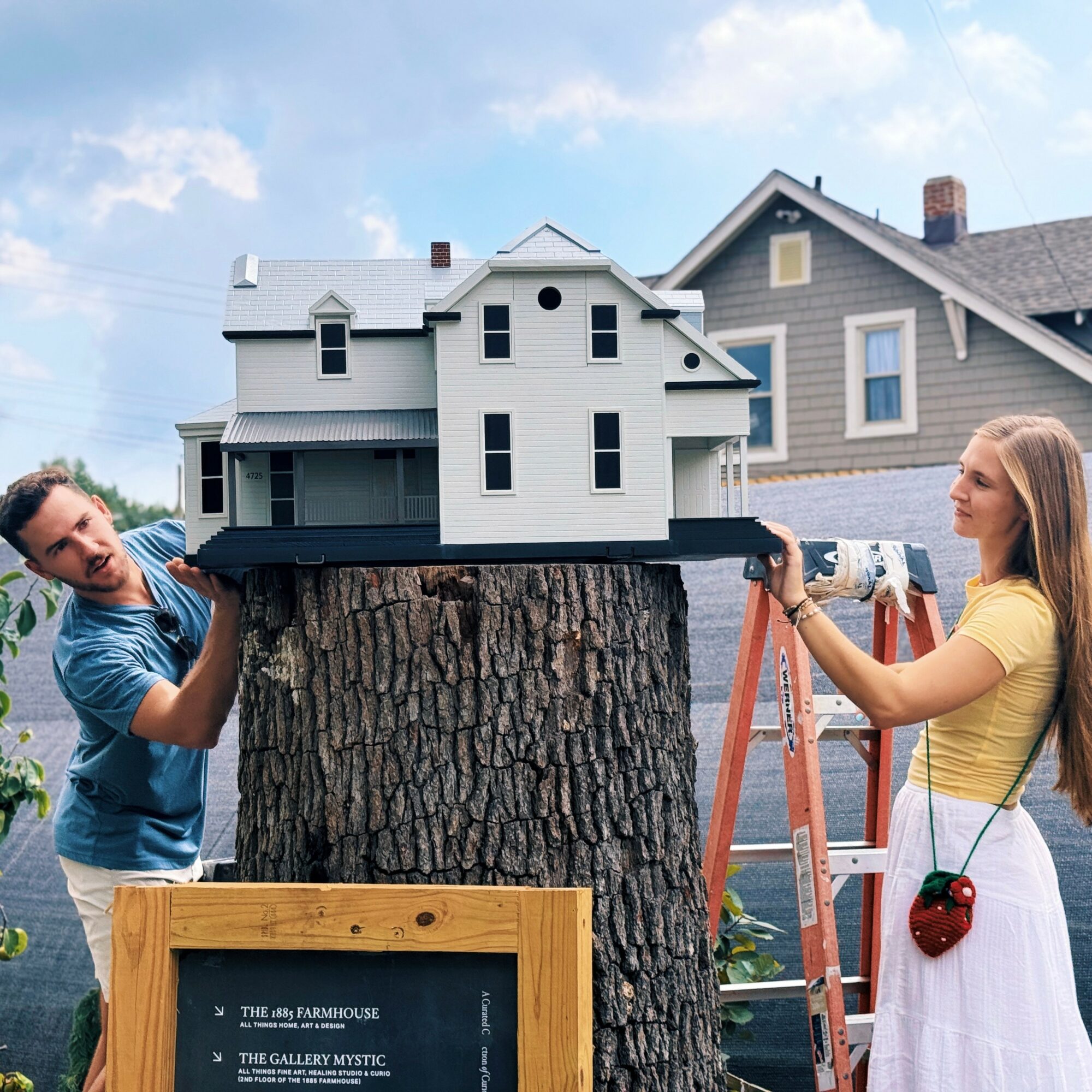 Two people work on a miniature house model on a tree stump, with a ladder and houses in the background.
