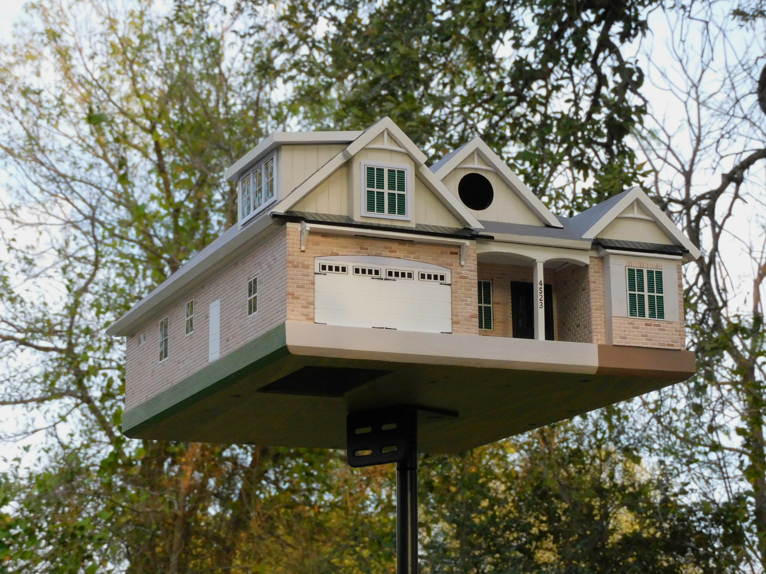 A house-shaped birdhouse mounted on a pole, with trees and sky in the background.