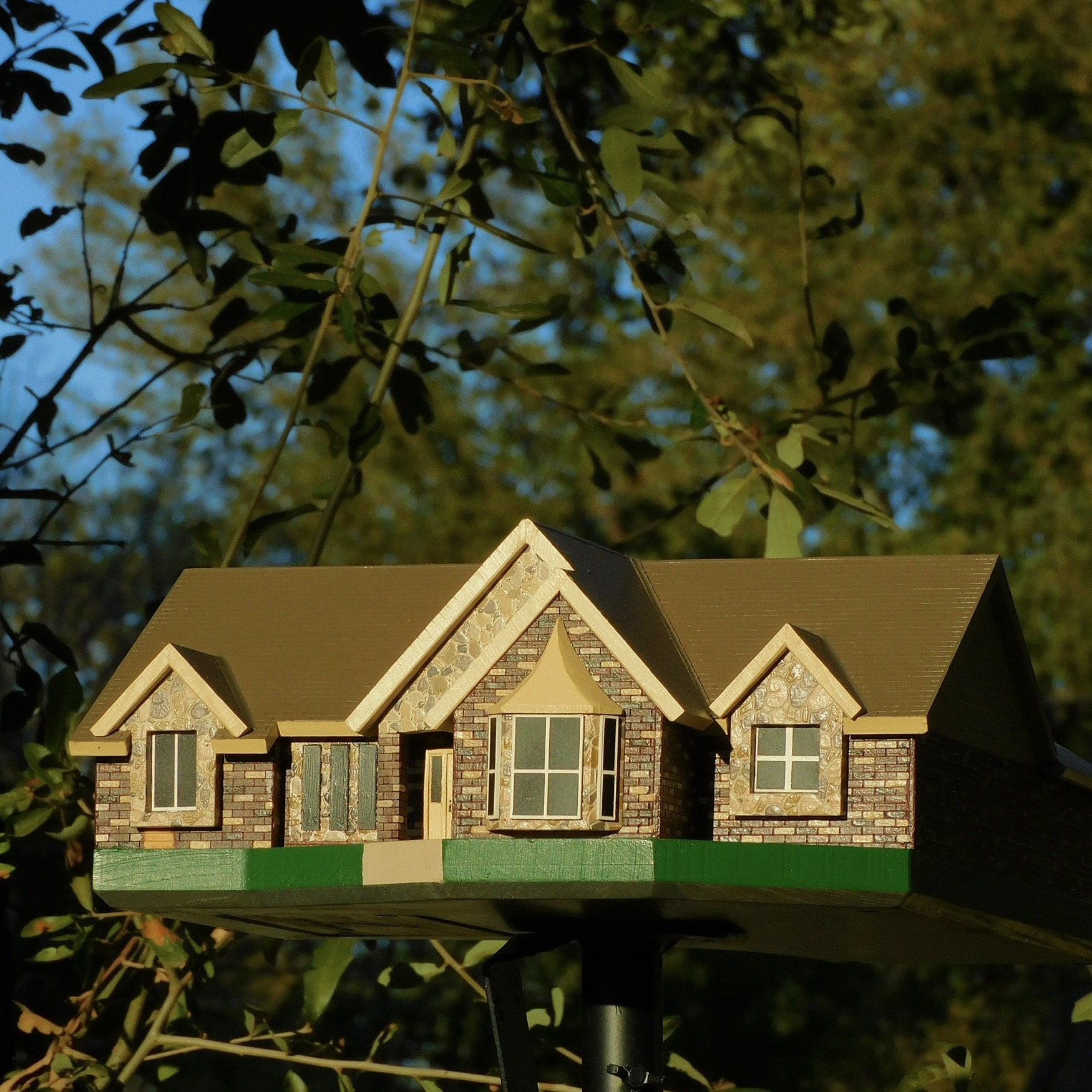 Model house with brick walls and multiple gabled roofs, set outdoors with trees and sky in background.