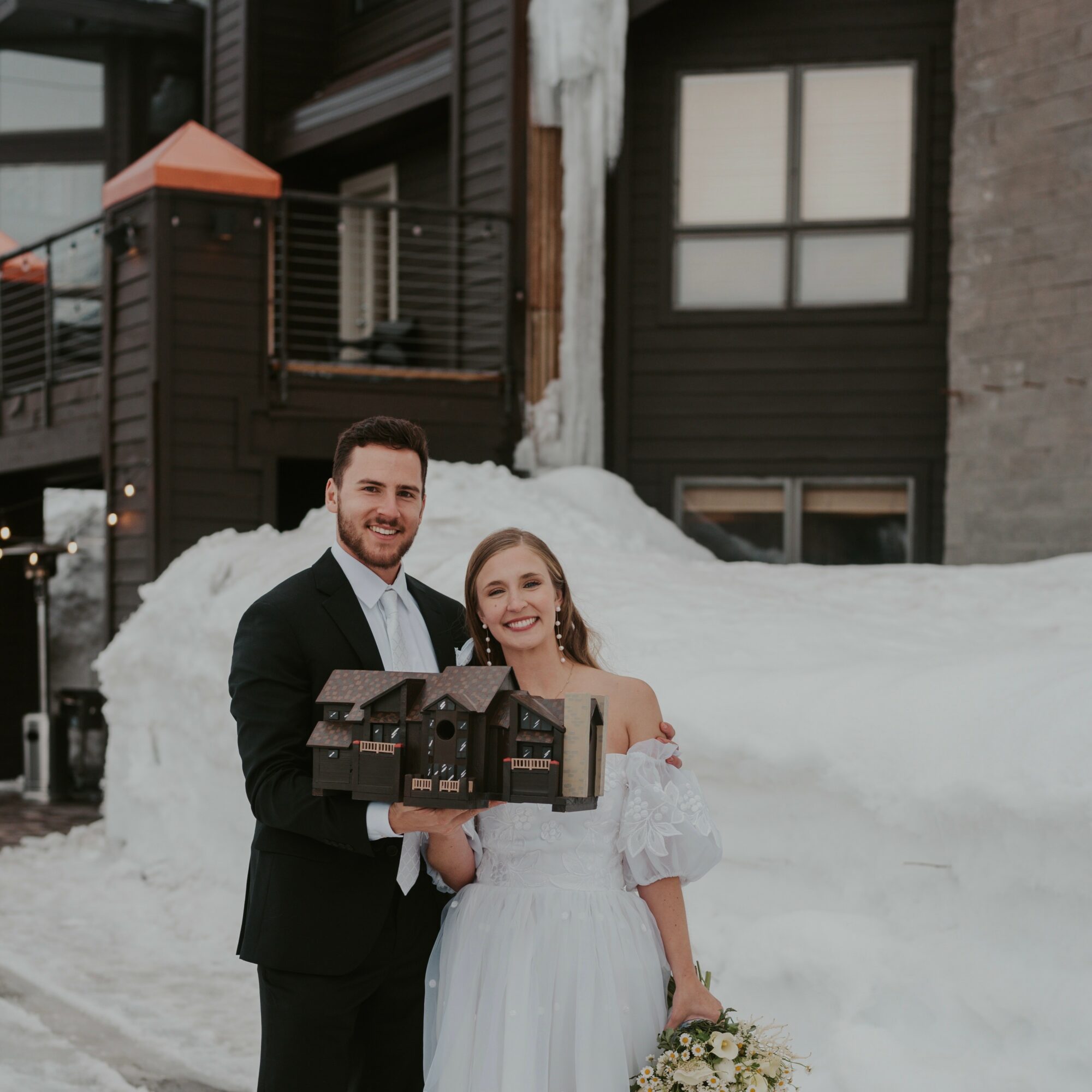 A smiling couple holding a model house outdoors in snowy setting, with a modern building in background.