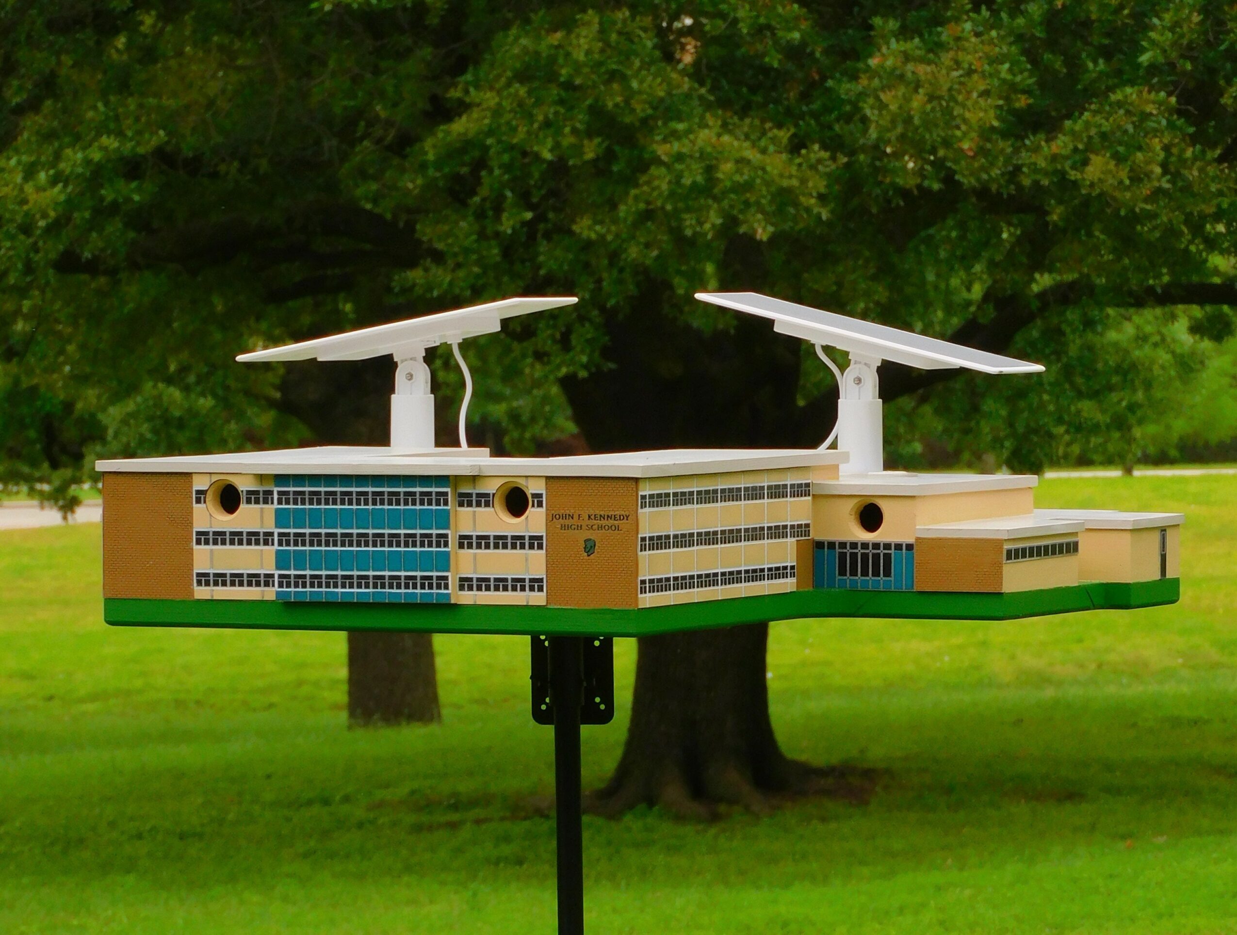 Model of a large building with two solar panels on top, set outdoors with trees and grass in the background.