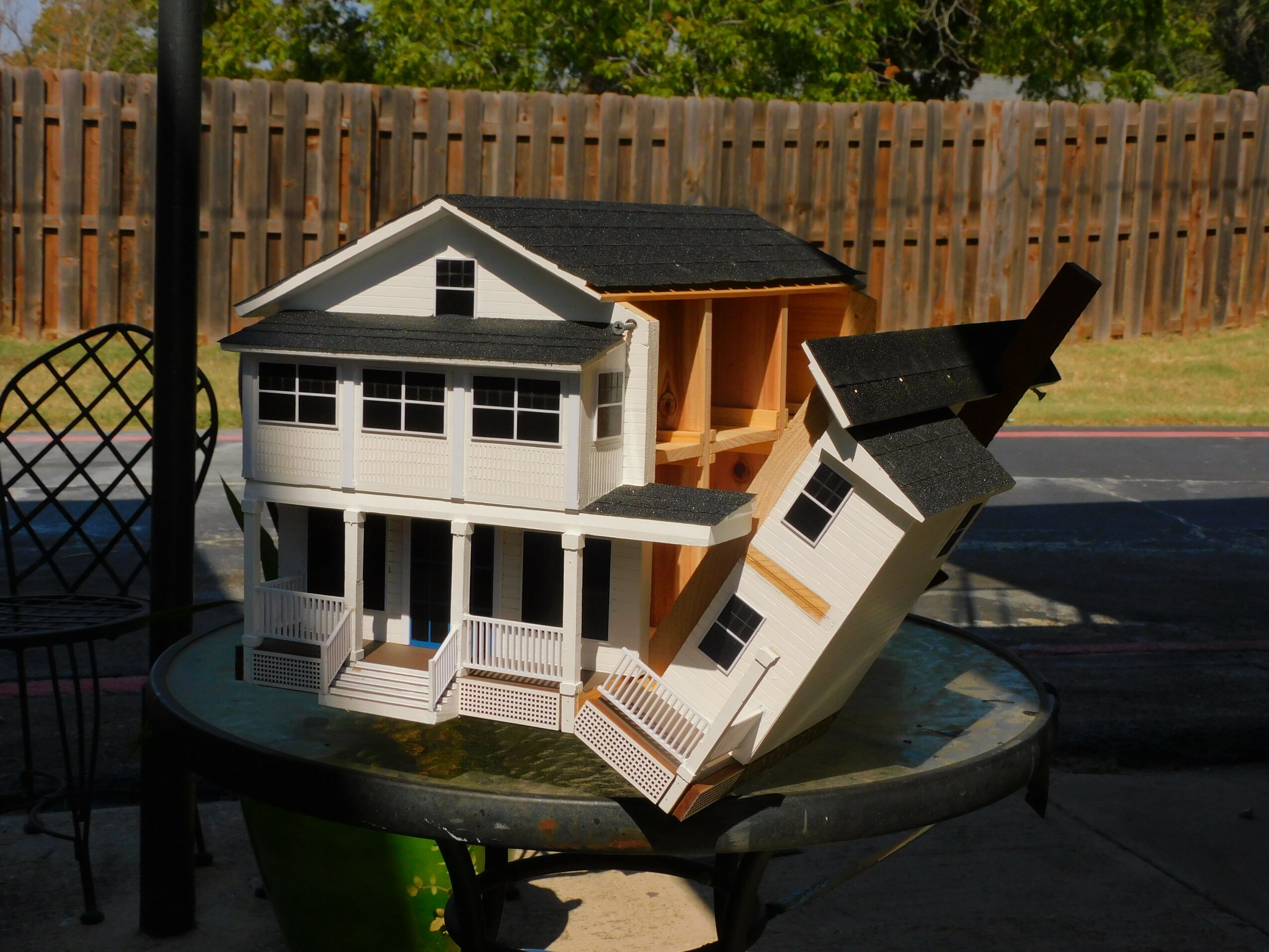 Model house with multiple levels and a sloped roof, placed on a round table outdoors, with a wooden fence in background.