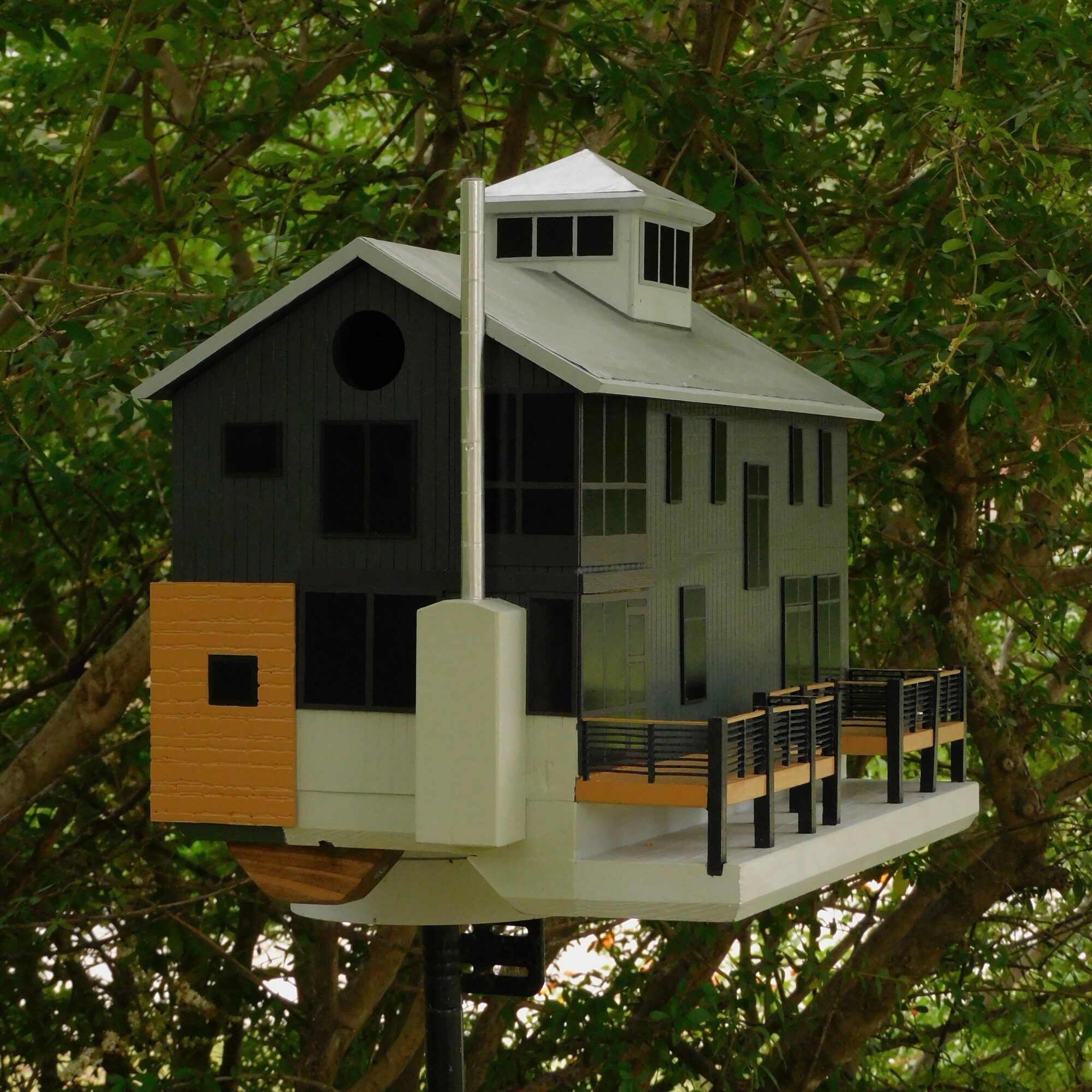Model house with black, white, and yellow sections, multiple windows, and a small porch, set against green foliage.