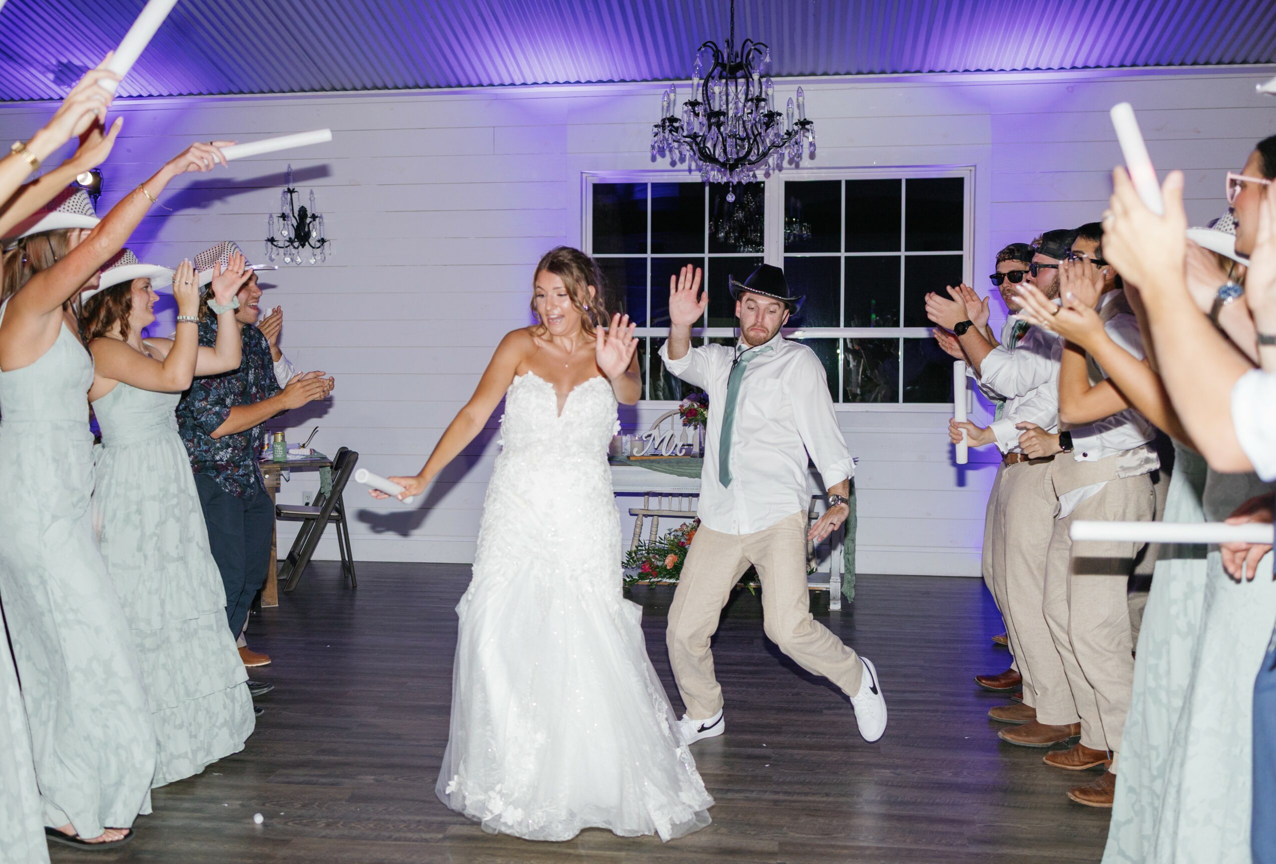Bride and groom dancing with guests clapping and celebrating indoors, chandelier hanging from ceiling, large window in background.