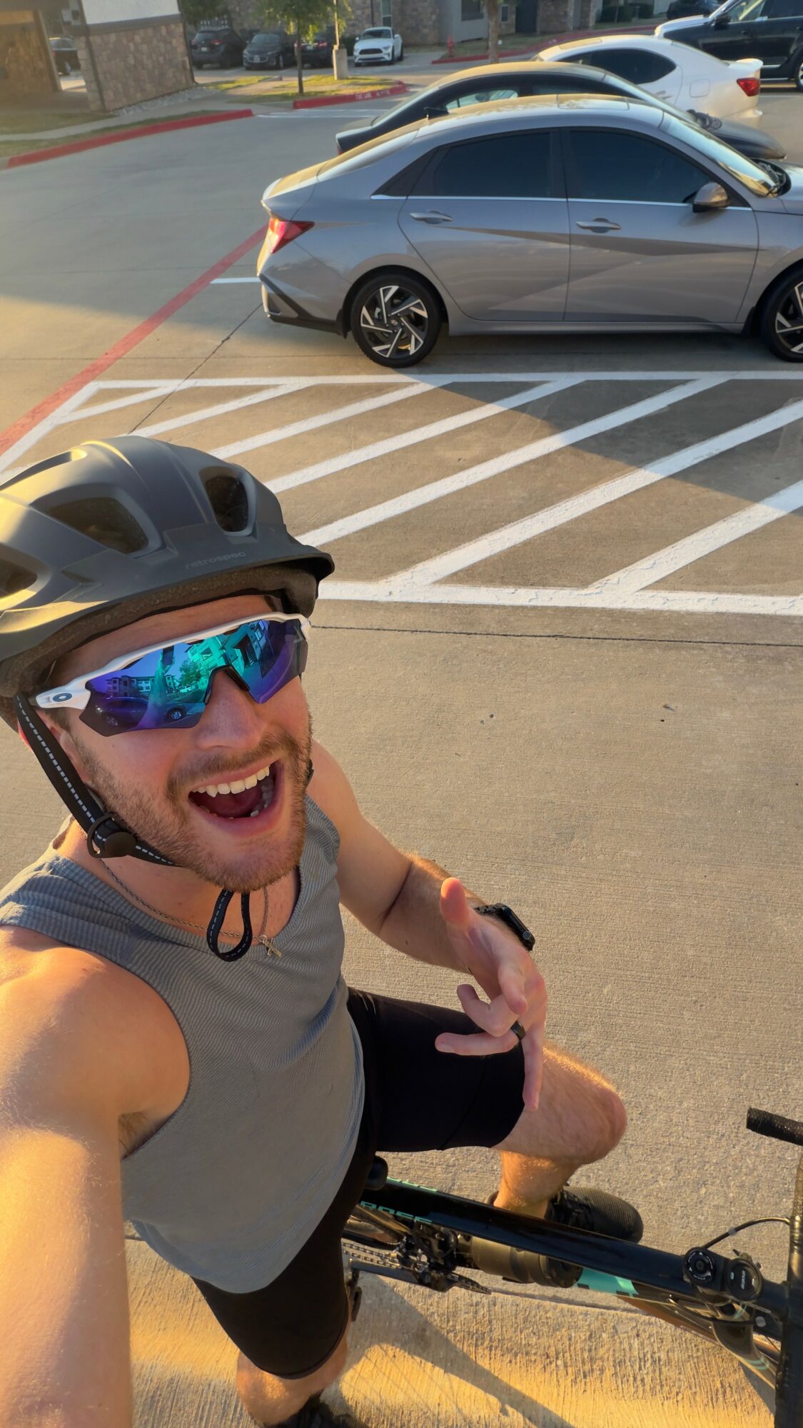 Man wearing helmet and sunglasses taking a selfie on a bicycle in a parking lot with cars behind him.