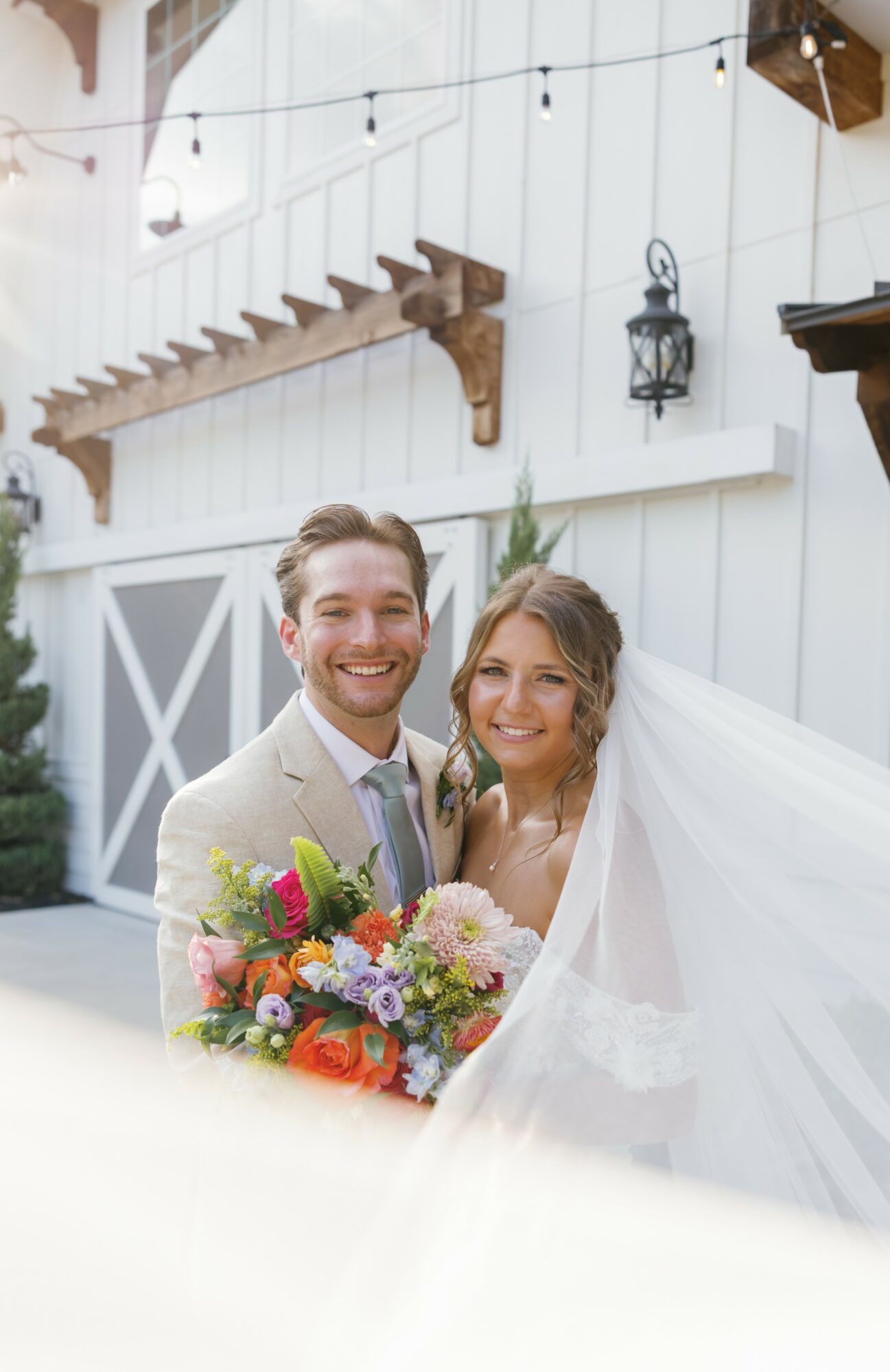 A smiling bride and groom holding a colorful bouquet outdoors, with a white wooden building and decorative lanterns in the background.
