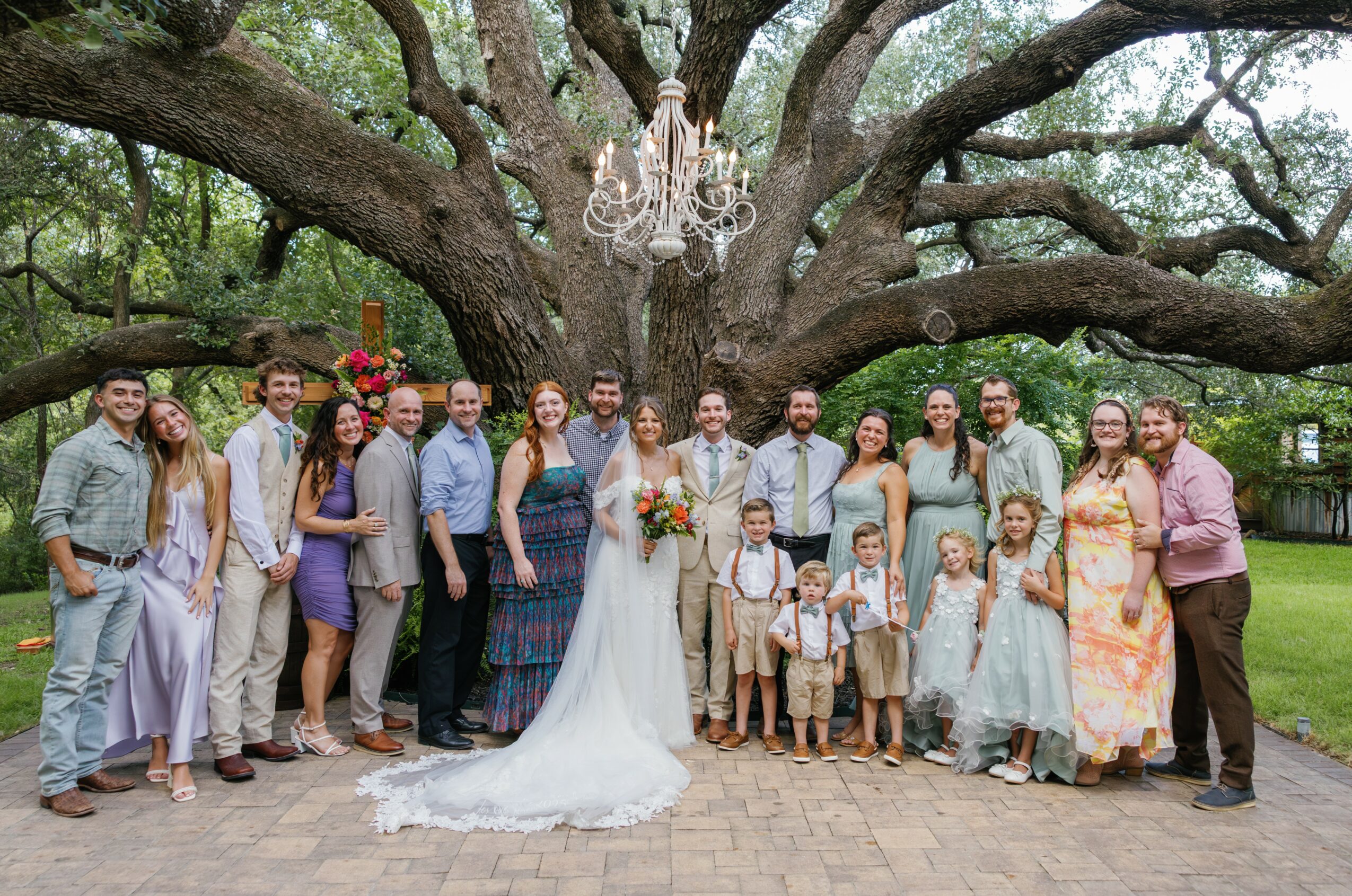 Group of people, including a bride and groom, standing outdoors under a large tree with a chandelier hanging from it.