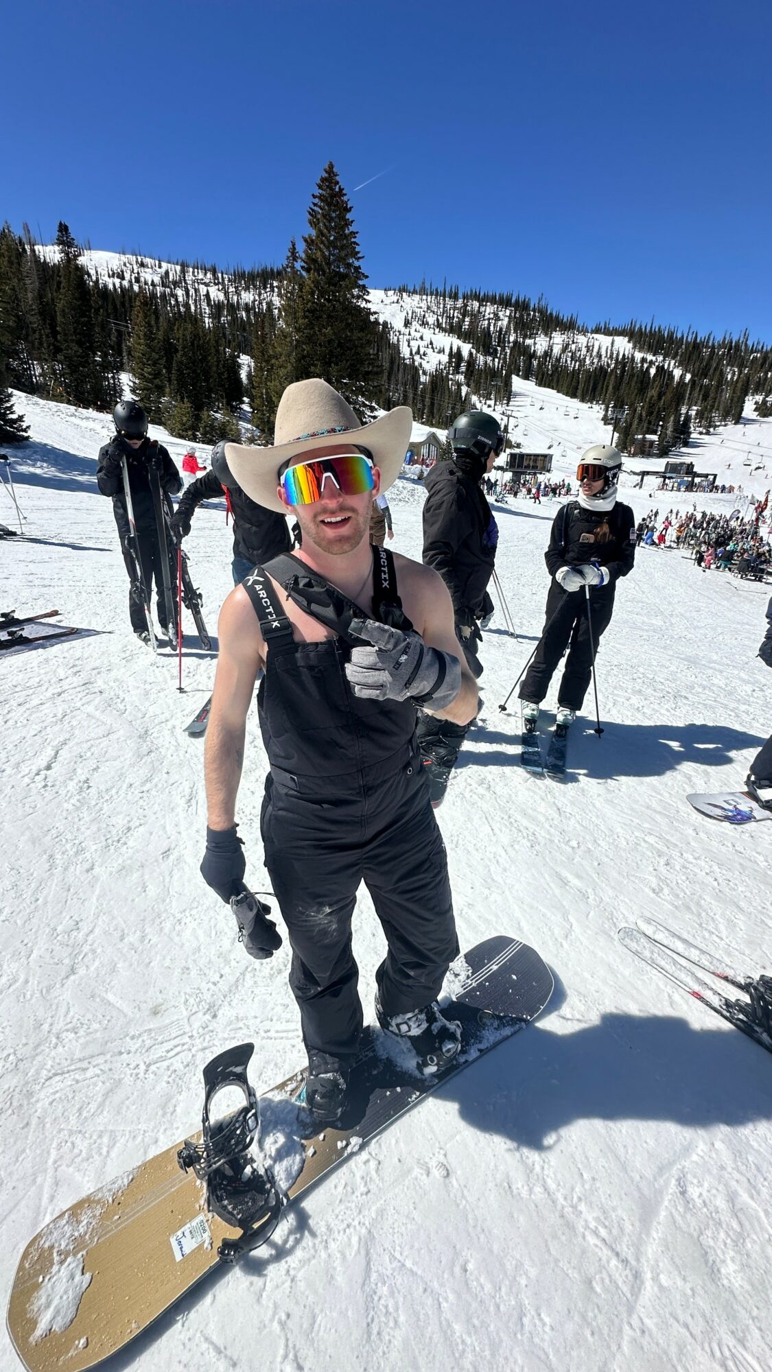 Person wearing a large hat and sunglasses on a snowboard in a snowy mountain landscape with trees and other people in the background.