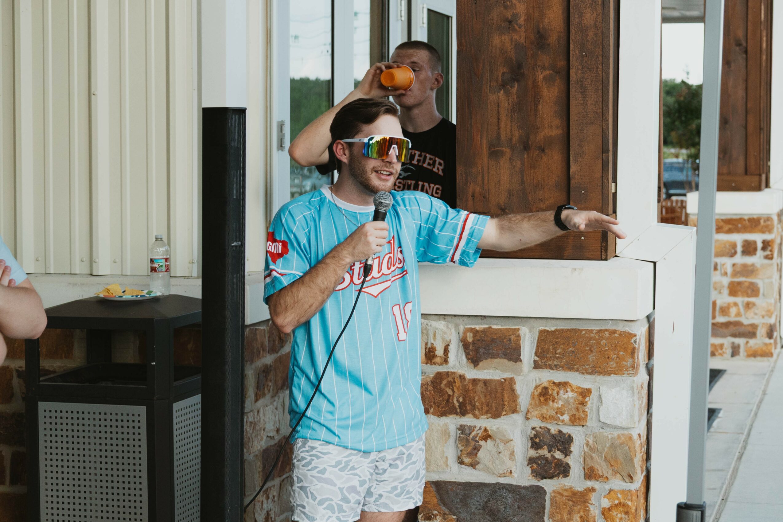 Person wearing sunglasses and a blue sports jersey holding a microphone, standing outside near a brick wall.