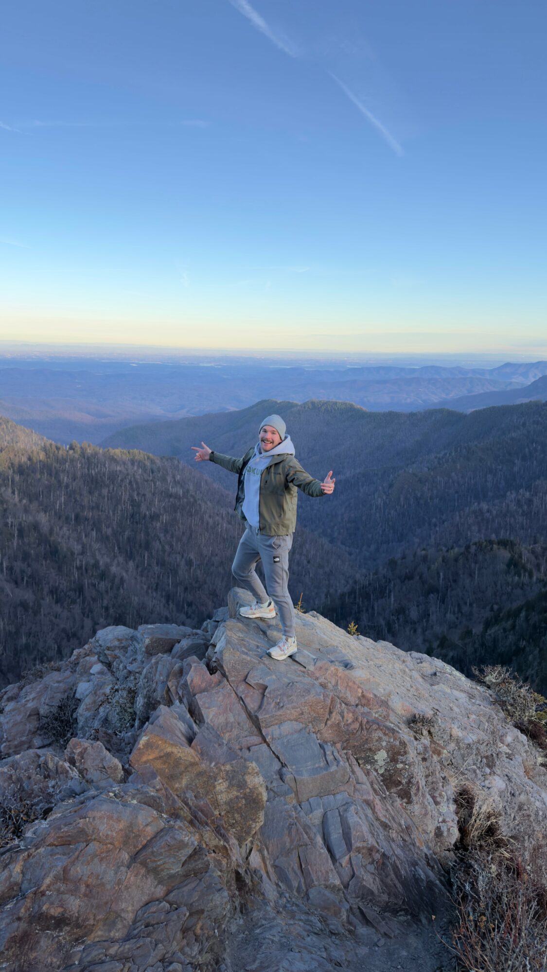 Person standing on rocks with arms outstretched on mountain peak, overlooking a vast landscape with mountains and sky.